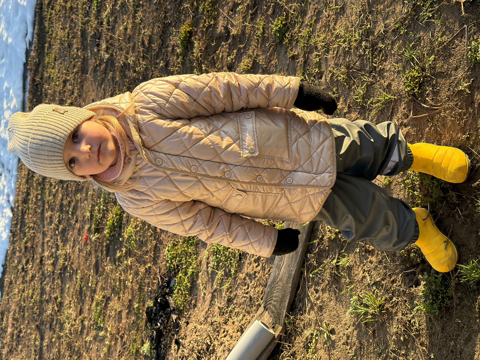 A young child stands in a field of dirt and sparse grass, dressed in a tan, quilted jacket, gray pants, black gloves, a gray knit hat, and yellow rubber boots. Patches of snow are visible in the upper left corner. A wooden plank and gray pipe lie on the ground in the foreground. The location is near Chaikovsky, Russia.
