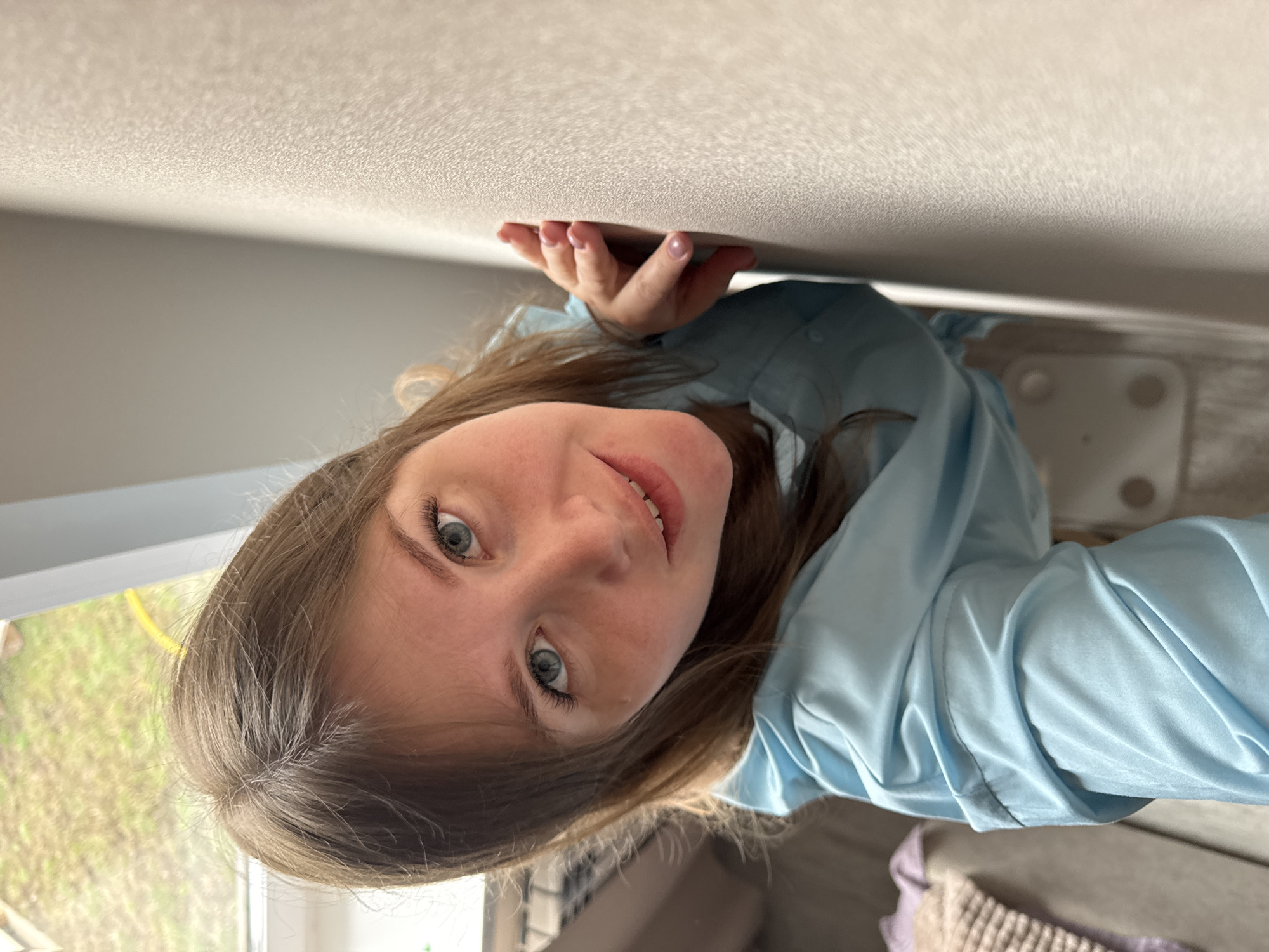 A young woman with long, light brown hair and blue eyes is playfully captured in an inverted perspective, appearing to look down from a ceiling. She has a subtle smile and is looking directly at the camera. Her right hand, with light pink painted fingernails, is resting against the textured white surface above her, enhancing the illusion of her being upside down. She is wearing a light blue, long-sleeved shirt or robe.
In the upper background (which would be the floor if the image were rotated), there's an indoor setting with a blurred white object with circular indentations, possibly a bathroom scale, and a soft, light brown knitted blanket or textile on a surface. To the left, a window provides a view of a lush green outdoor area, with what appears to be a yellow hose or pipe visible. The scene is brightly lit, suggesting it's daytime, likely using natural light from the window. The overall impression is one of a playful portrait taken indoors, potentially in a home in Ферма, Russia, with an unconventional photographic angle creating an engaging visual effect. There is no visible text.