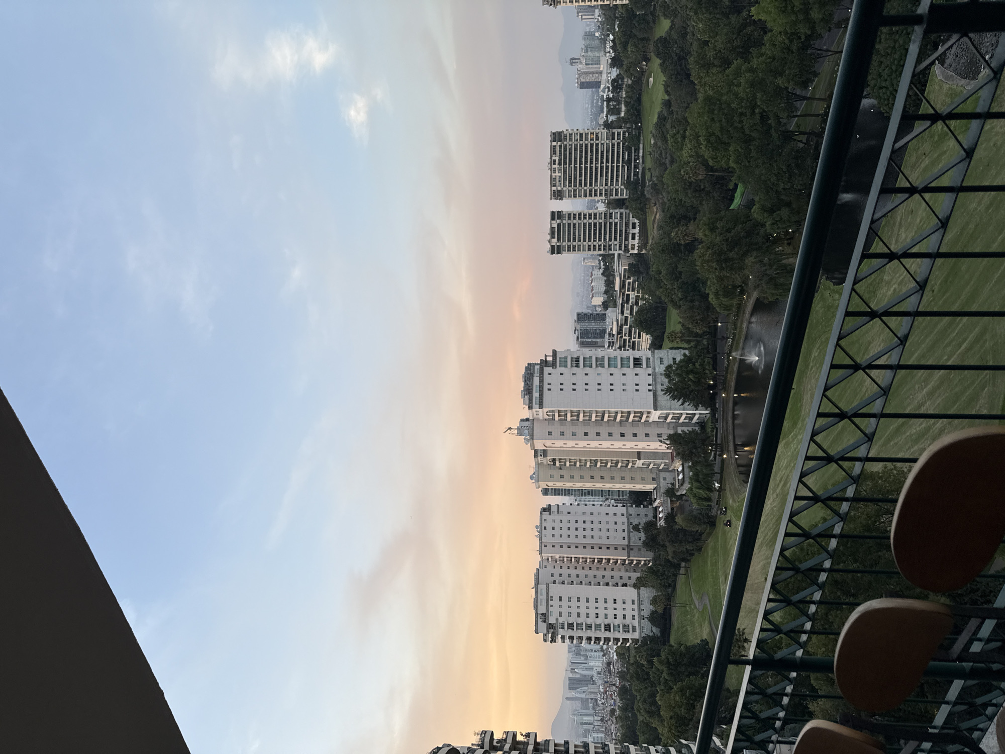 The image presents a wide, elevated view of an urban landscape, likely taken from a balcony during the golden hour, possibly sunset, given the sky's colors. In the immediate foreground on the right, a dark metal balcony railing with a patterned design is prominent, along with parts of two light brown, possibly wooden, chairs.
Beyond the balcony, the midground features a sprawling, manicured green space, likely a golf course or a large park, adorned with numerous trees and a winding path. A small body of water, possibly a pond or lake, is visible within this green area, complete with a fountain spraying water. Several modern, multi-story buildings with light-colored facades and numerous windows line the edges of this green expanse, forming a significant part of the cityscape.
In the background, the city extends further with more buildings fading into a hazy urban sprawl, and distant mountains are faintly visible on the horizon. The sky transitions from a soft, light blue with wispy clouds on the left to warm hues of orange and yellow on the right, suggesting the early evening light. The overall scene depicts a serene urban environment with a blend of natural green spaces and modern architecture. There is no visible text in the image. The setting suggests a large metropolitan area, consistent with Mexico City.