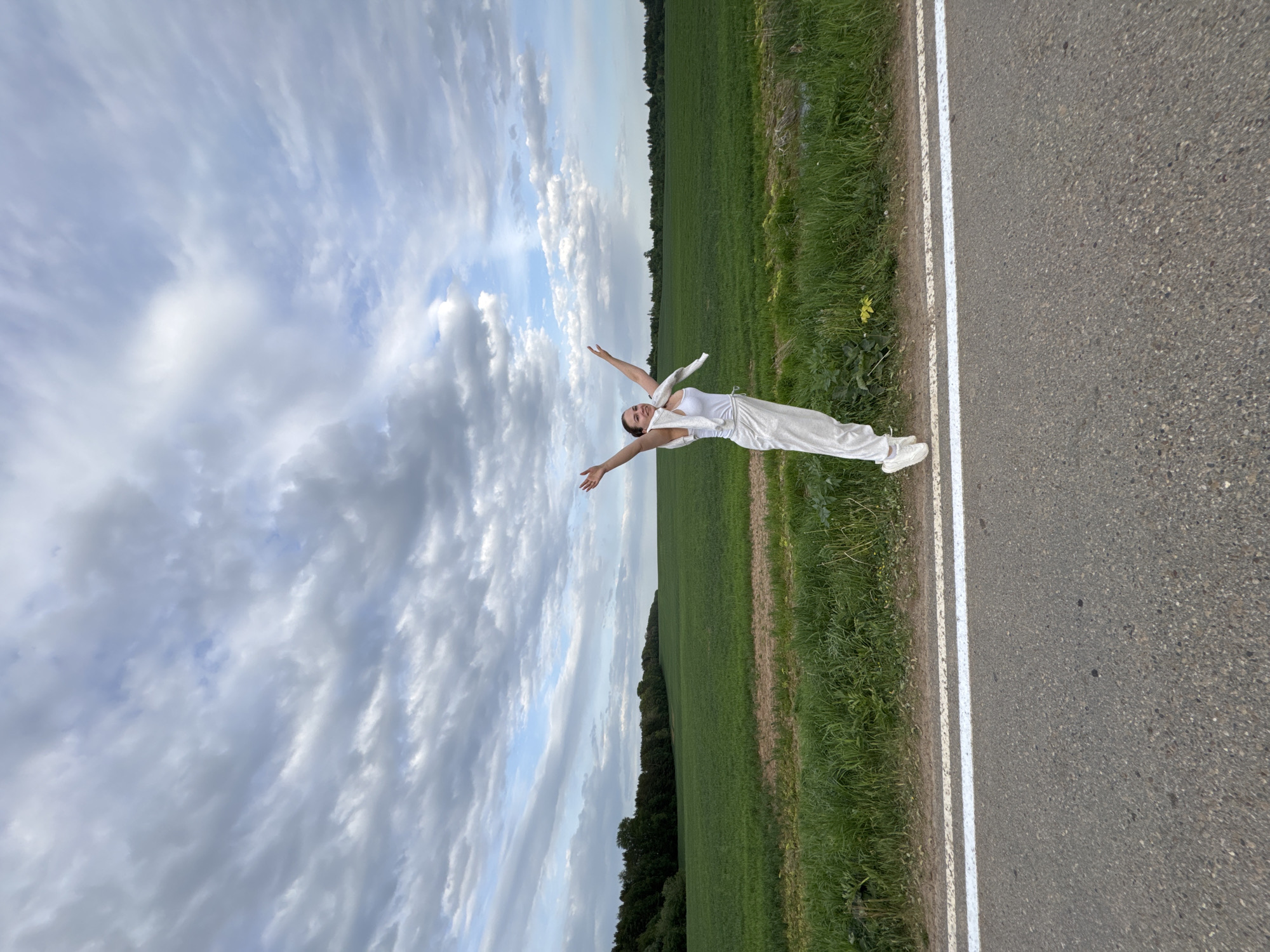 A woman in a white outfit stands with her arms outstretched on the side of an asphalt road in a rural area. A field of green grass stretches beyond the road. The sky overhead is partly cloudy. There are trees in the distance. The location is in the vicinity of Lukintsy, Russia.