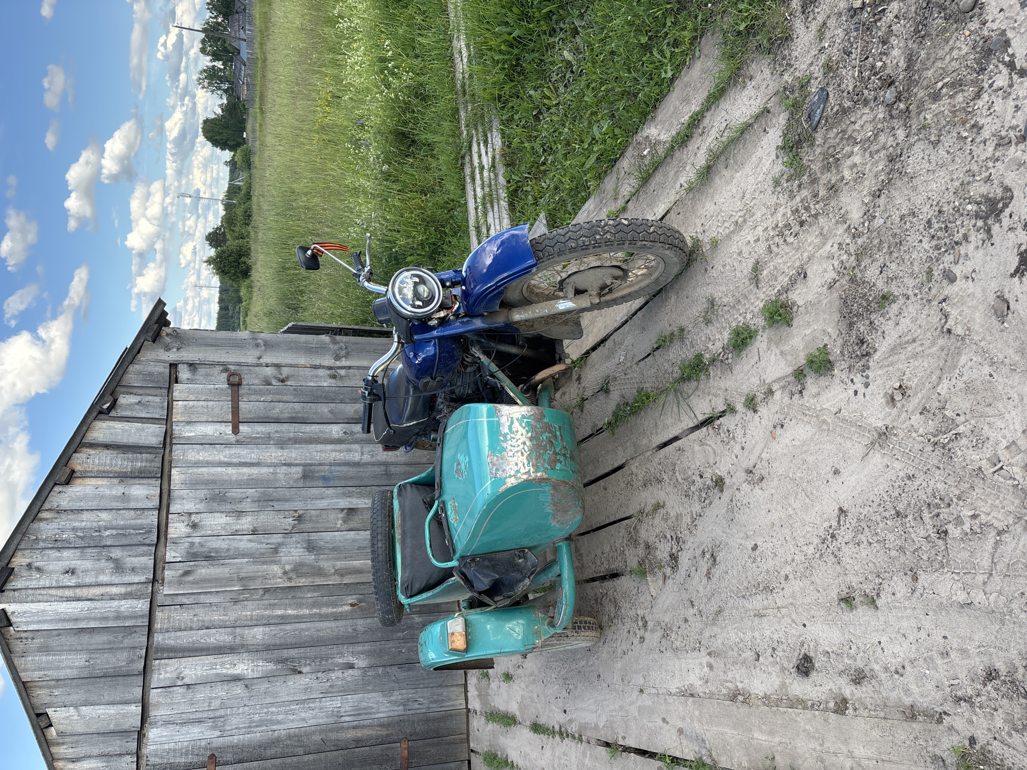 A blue motorcycle with a turquoise sidecar is parked on a weathered wooden platform next to a rustic wooden shed. The motorcycle's headlight is round and silver, and it has chrome handlebars. A red and orange ribbon is attached to the left handlebar. The sidecar appears to be slightly dented and has some chipped paint, revealing a metallic silver underneath. The platform is made of rough wooden planks, and there are tire tracks in the dirt and gravel surrounding it.
Behind the shed, a field of tall green grass stretches towards a distant treeline. The sky is a bright blue with fluffy white clouds. In the background, there are some structures visible, including what appears to be a cell tower and a few buildings that suggest a rural settlement.
The scene is outdoors, likely in a rural area of Kurgan, Russia. The bright sky and abundant greenery suggest it is daytime, possibly late morning or early afternoon, under fair weather conditions. There is no activity or event taking place; the image appears to be a still capture of the parked motorcycle and its surroundings. The overall impression is one of a quiet, perhaps somewhat neglected, rural setting. No visible text is present in the image.