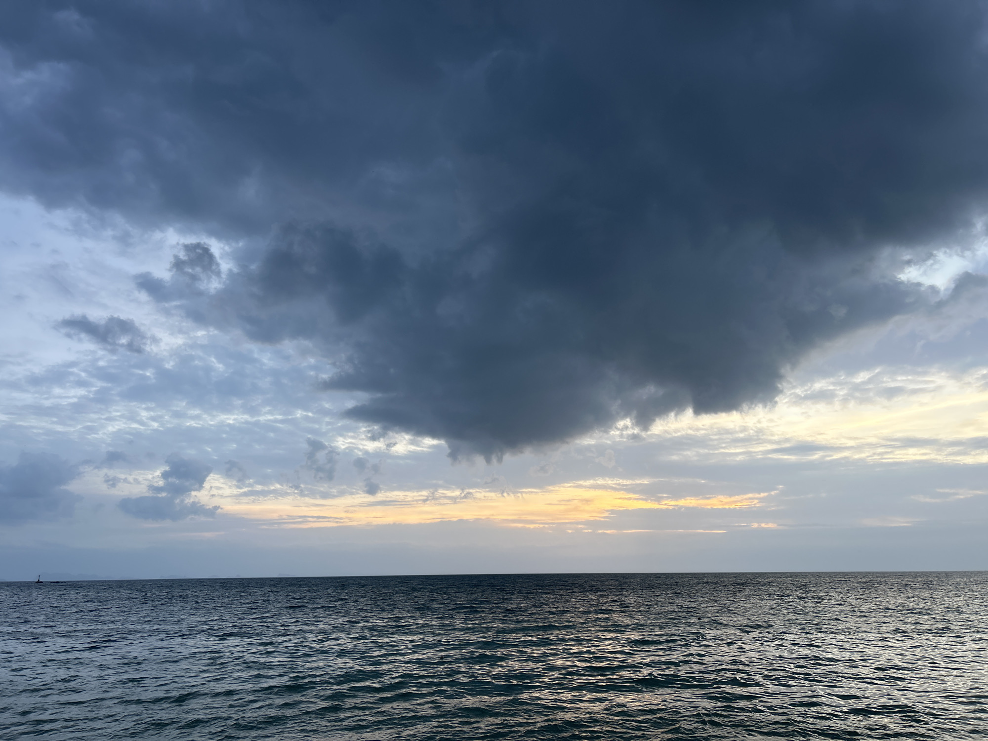 An outdoor, eye-level, wide shot captures the horizon of a vast, dark ocean under a cloudy sky at dusk. The water exhibits subtle ripples, reflecting the dim light. Dominating the upper portion of the frame are heavy, dark grey clouds, interspersed with patches of lighter, translucent clouds and streaks of pale yellow and orange light near the horizon, indicative of the setting sun. In the far distance, a small, dark object, possibly a buoy or a distant structure, is visible on the left. The scene evokes a sense of calm before a potential storm or the transition from day to night. The location is the Ko Pha-ngan District in Thailand.