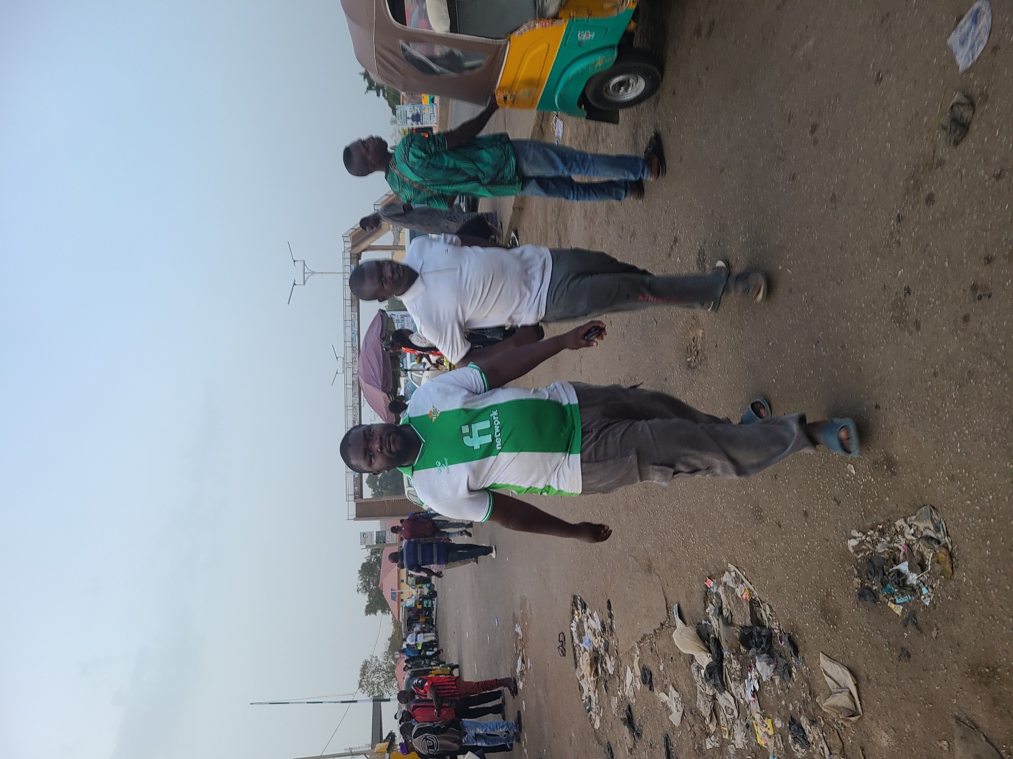 The image shows an outdoor scene in what appears to be a roadside area in Jos, Nigeria. Three men are prominently featured in the foreground. The first man is wearing a white and green soccer jersey with a "Network" logo. The second man is wearing a white polo shirt, and the third man is wearing a green patterned shirt and jeans. All three men are dark-skinned. In the background, there is a crowd of people, most of whom are also dark-skinned. There is a three-wheeled vehicle. There is a significant amount of litter scattered on the ground. Overhead, there is a lightly overcast sky. Streetlights and other structures are visible in the background.