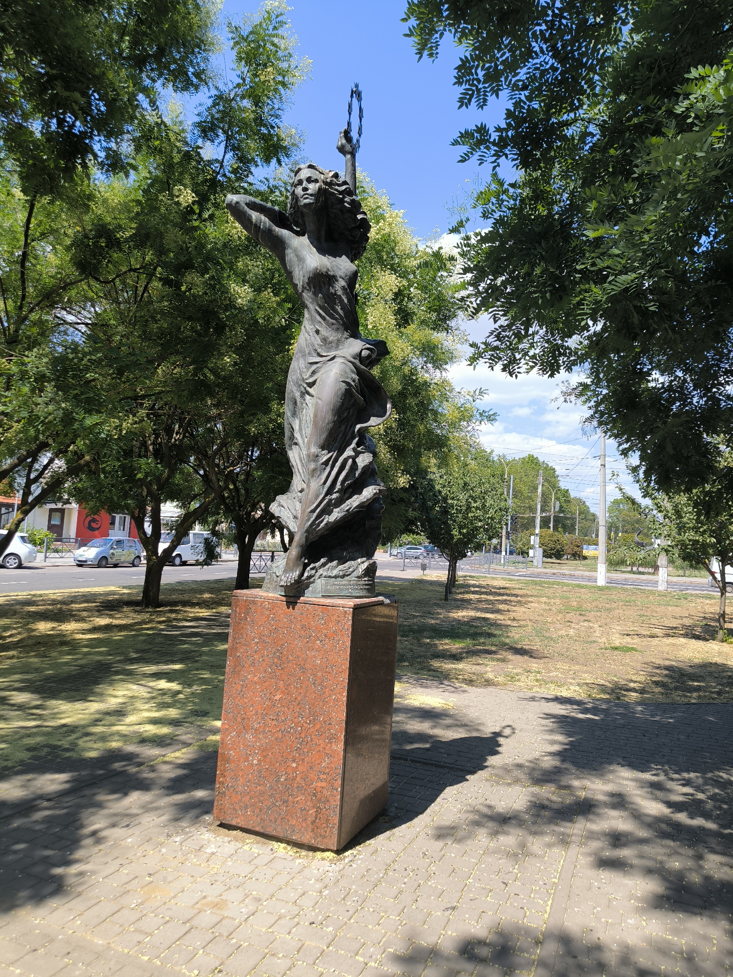 A bronze statue of a woman stands on a red granite pedestal in a park-like setting in Mykolaiv, Ukraine. The statue depicts a woman with her arms raised, holding a laurel wreath or similar object aloft. She is dressed in flowing robes that appear to be windblown. The statue is situated on a paved walkway, with a grassy area behind it. In the background, there is a street with cars, trees, and utility poles. The sky is blue with some scattered clouds. The statue is casting a shadow on the walkway. The overall scene suggests a public monument in an urban environment, likely a park or plaza.