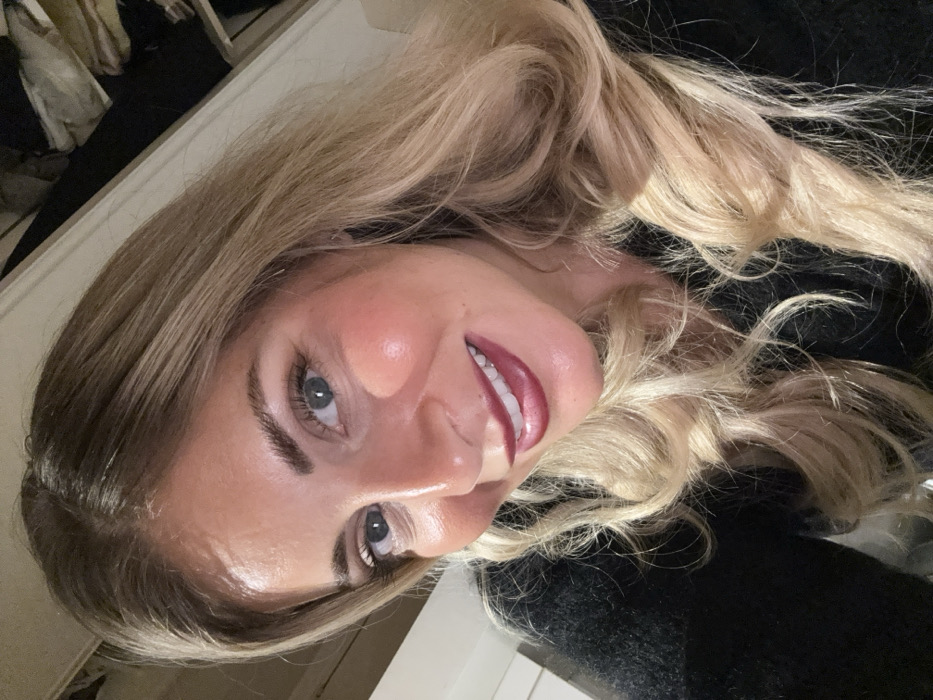 A woman poses, her face angled toward the camera. Her hair, a mix of blonde and brown, is styled with waves and movement. She wears lipstick in a deep berry shade. Her skin has a noticeable glow, and her eyes are light-colored, framed by dark eyebrows. A dark-colored garment is visible behind her. The background has hints of a room setting.