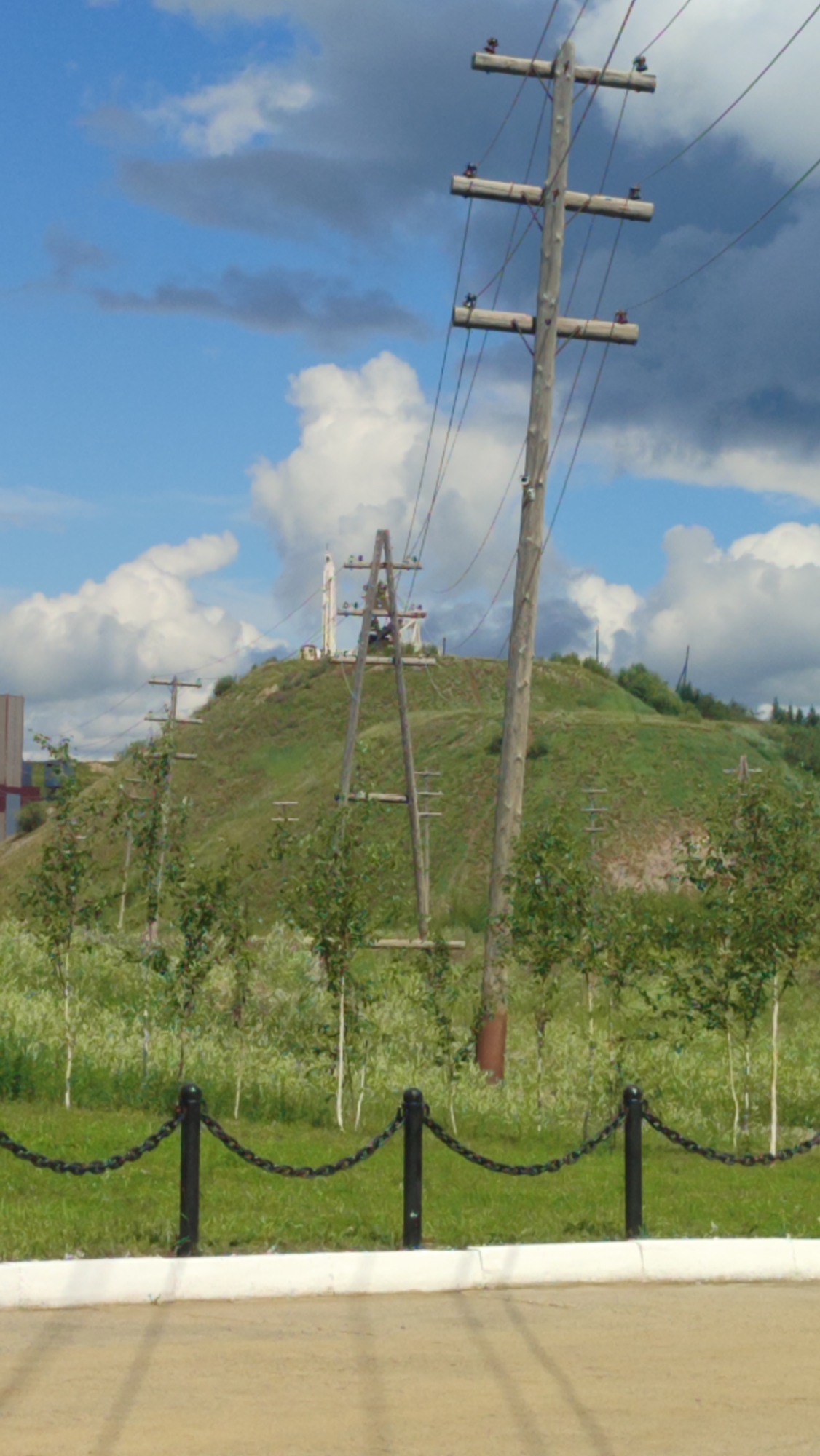 The image shows a landscape scene with a prominent wooden utility pole in the foreground, supporting multiple wires. Behind it, a grassy hill rises, dotted with trees and other utility poles. In the distance, a tall, white structure, possibly a communication tower or antenna, is visible on top of the hill. The sky is partly cloudy with a mix of blue and dark gray clouds. The foreground features a paved area and a black chain-link fence. The scene is likely located in Mirny, Russia, given the presence of utility infrastructure and the general landscape. There are no people or animals visible in the image.