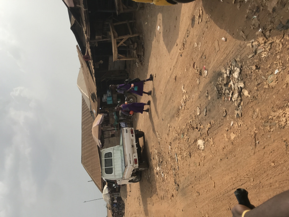 The image depicts a bustling street scene in Ogbese, Nigeria, under an overcast sky. A wide, unpaved dirt road, littered with debris and tire tracks, dominates the foreground and mid-ground.
In the mid-ground, a white compact pickup truck with a light blue bed liner is parked facing away from the viewer on the left side of the road. Two young females, likely schoolgirls, wearing matching purple uniforms with black shoes and socks, walk from left to right across the road in front of the truck. The girl on the left carries an orange item, and the girl on the right carries a white or light-colored container or bag. They cast discernible shadows to their left.
To the right of the walking girls, the edge of a yellow vehicle is visible. Along both sides of the road, particularly in the background, numerous market stalls and single-story structures with corrugated metal roofs are present. Several stalls feature large shade umbrellas, one tan and one white. Visible items in the market include baskets, crates, and wooden tables used for display. Further down the road on the left, other individuals and motorcycles are present near additional market activity. A bare arm with a yellow band is partially visible in the bottom left corner of the frame.