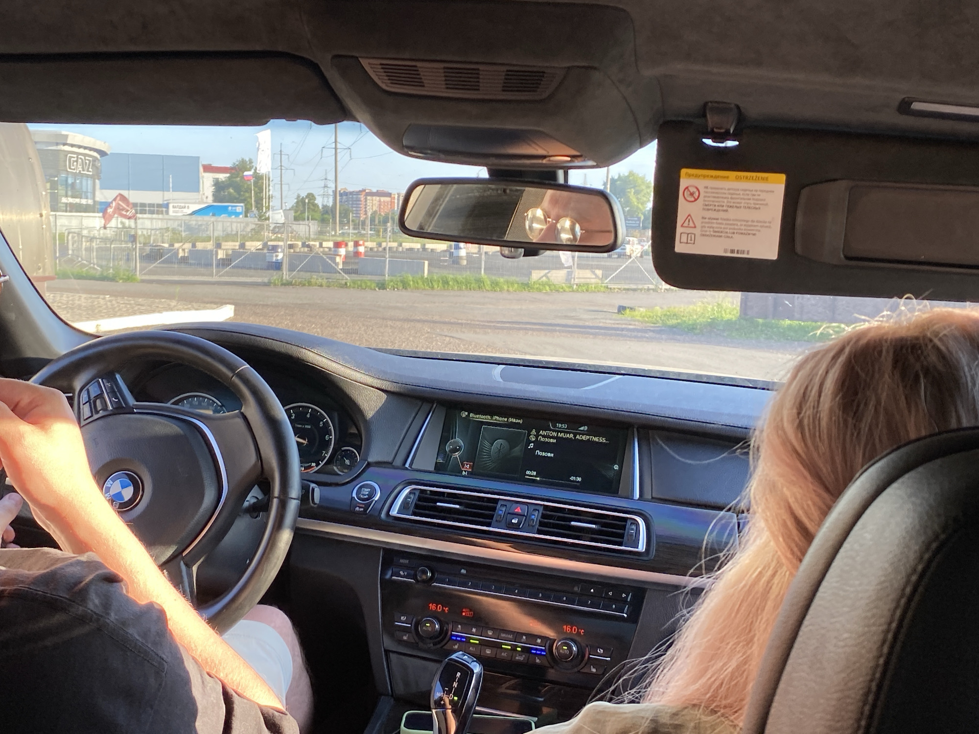 The image shows the interior of a car, looking forward through the windshield. A person's arm and hand, partially obscured by sunlight, are visible on the left side, resting on the steering wheel. The steering wheel is black with a BMW logo in the center. The dashboard includes a central infotainment screen displaying music information and a clock. Below the screen are climate control buttons and a gear shift lever. To the right, the side of a person's head with long, light-colored hair is visible, suggesting they are a passenger. The exterior view through the windshield shows buildings, power lines, and a fenced area with construction barriers, indicating an urban or industrial setting. A sun visor with a warning label is visible at the top of the windshield. The overall scene depicts a car journey in a city.