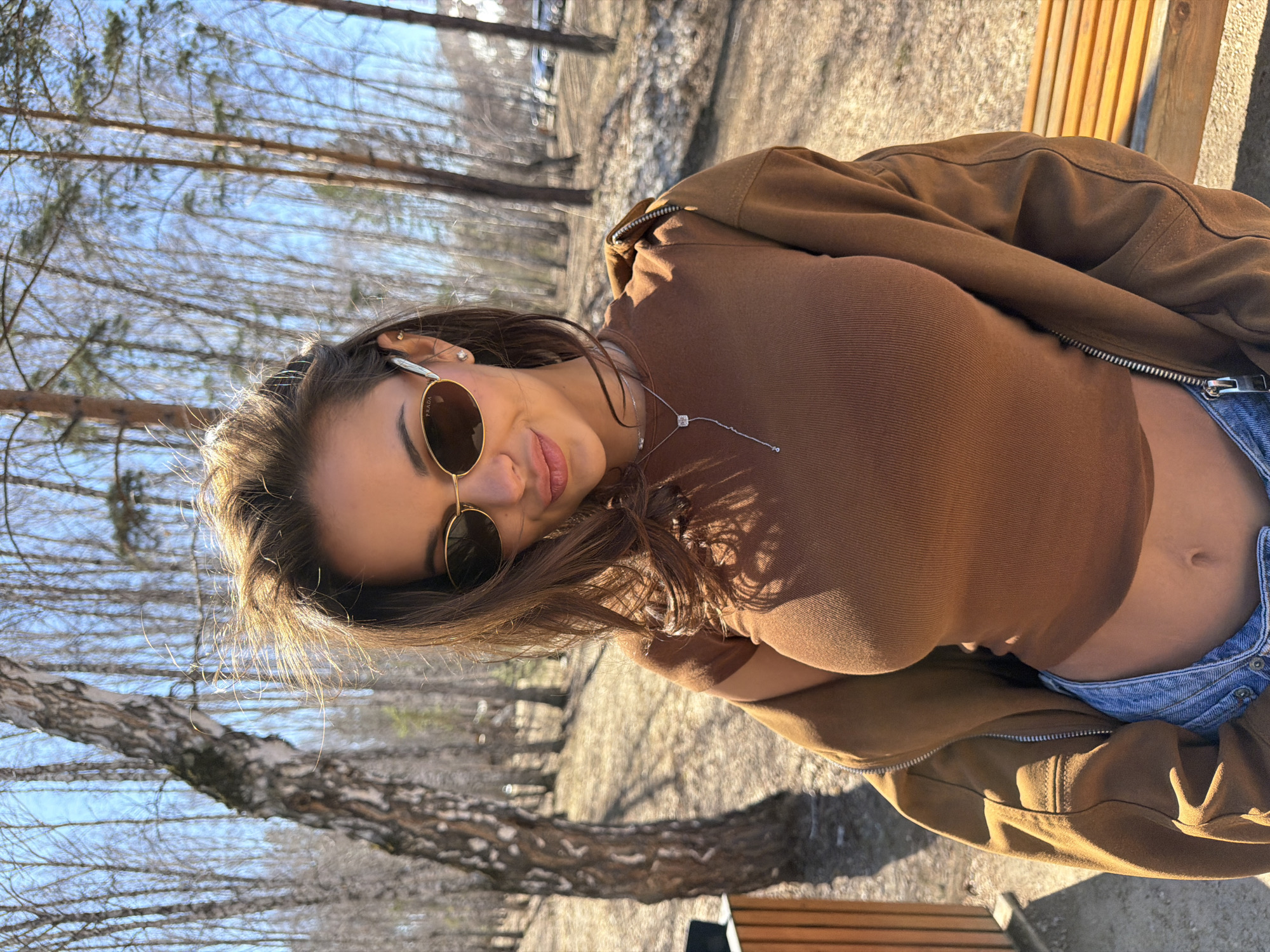 A young woman is captured in a sunlit outdoor portrait, appearing to be sitting or leaning on a wooden bench in a park or forest, likely in Krasnoyarsk, Russia. She looks directly at the camera with a relaxed, subtle smile or smirk, her head tilted slightly.
She has long, dark brown hair and is wearing round, gold-rimmed sunglasses, with the brand "PRADA" faintly visible on the right temple. Her attire includes a form-fitting, ribbed brown long-sleeved top and a brown zip-up jacket that is partially unzipped, revealing her midriff and the light blue denim waistline of her bottoms. She accessorizes with small stud earrings and a delicate silver necklace featuring a square pendant.
The background is composed of numerous bare-branched trees, suggesting a late winter or early spring setting. The bright sunlight casts clear shadows, indicating a sunny day. The ground beneath the trees appears to be dirt or gravel. The scene conveys a casual and bright atmosphere.