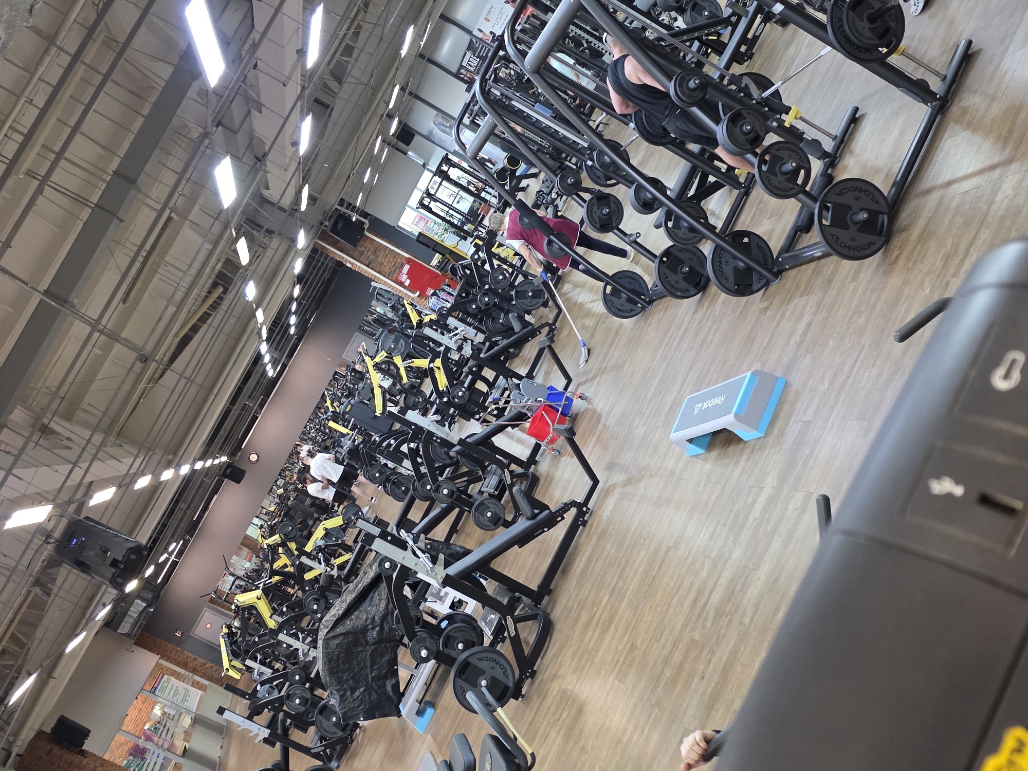 Here is a description of the image:
**Scene:** The image is an interior shot of a fitness center or gym, likely located in Chaikovsky, Russia based on textual clues in the image.
**Environment:** The gym is well-lit with visible ceiling fixtures. Numerous exercise machines are visible in rows, including weight lifting stations. The floor is made of light wood.
**People:** There are several people present. One person is using a weight machine, another seems to be cleaning the floor with a long-handled broom.
**Objects:** The gym is filled with weightlifting equipment. There's a "Reebok" branded step platform near the machines. Weights with "TECHNOGYM" branding are visible. Also visible in the distance is a red container.
**Actions:** People are engaged in activities typical of a gym setting, like cleaning and exercising.
**Overall Impression:** It is a typical gym scene with people exercising and maintaining the space.