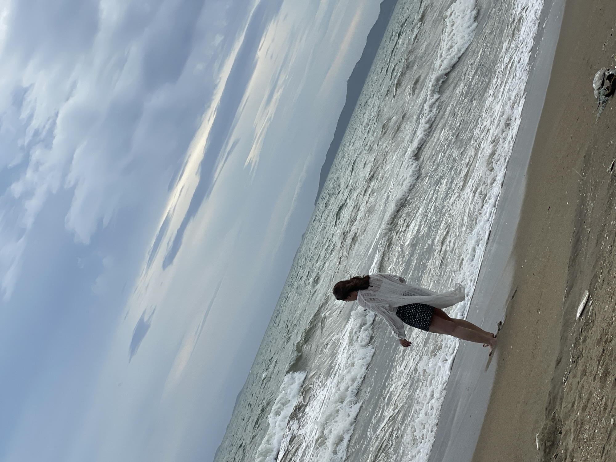 A lone woman stands on a sandy beach, facing the sea. The sky is overcast with clouds. The ocean is rough, with waves crashing on the shore. The woman is wearing a white cardigan over a black and white patterned dress. Her back is to the camera. In the background, a hilly coastline is visible. The scene suggests a cloudy day at the beach in Pattaya City, Thailand.