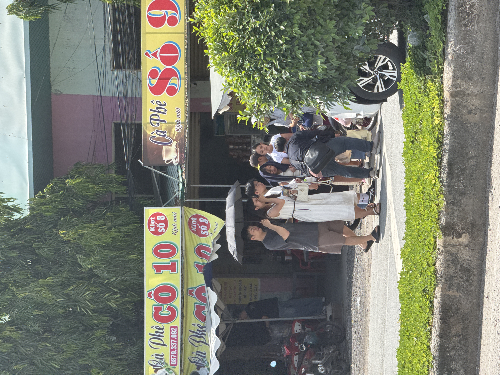 The image captures a street scene in Ben Cau, Vietnam. In the foreground, a patch of lush green ground cover borders a concrete curb and a grey asphalt road. A white car with distinctive silver and black alloy wheels is parked on the right side of the frame, partially obscured by a large, vibrant green bush.

On the street, several individuals are gathered. A group of people stand under a black umbrella, with one person holding it. A man in a dark t-shirt and brown shorts stands with black flip-flops. Next to him, a woman in a white dress and white sandals is holding a small white purse. Another man in a black shirt and dark pants is partially visible. To the left, a man in a grey t-shirt and jeans stands near a motorcycle.

In the background, two prominent yellow signs advertise "Cà Phê Cô 10" and "Cà Phê Số 9". The "Cà Phê Cô 10" sign features red lettering, the number "10" in red, and a phone number "0879.337.092". It also includes a smaller red circular sign with "KIOT SỐ 8" and "Kính mời". The "Cà Phê Số 9" sign also has red lettering and a depiction of a coffee cup. Behind the signs, a building with sections painted in pink and light blue is visible, along with electrical wires. An awning extends over part of the storefront area. A red motorcycle is parked to the left of the gathering. The overall environment suggests a commercial area, likely a street lined with shops or food stalls. The lighting indicates it is daytime, with bright sunlight creating shadows.