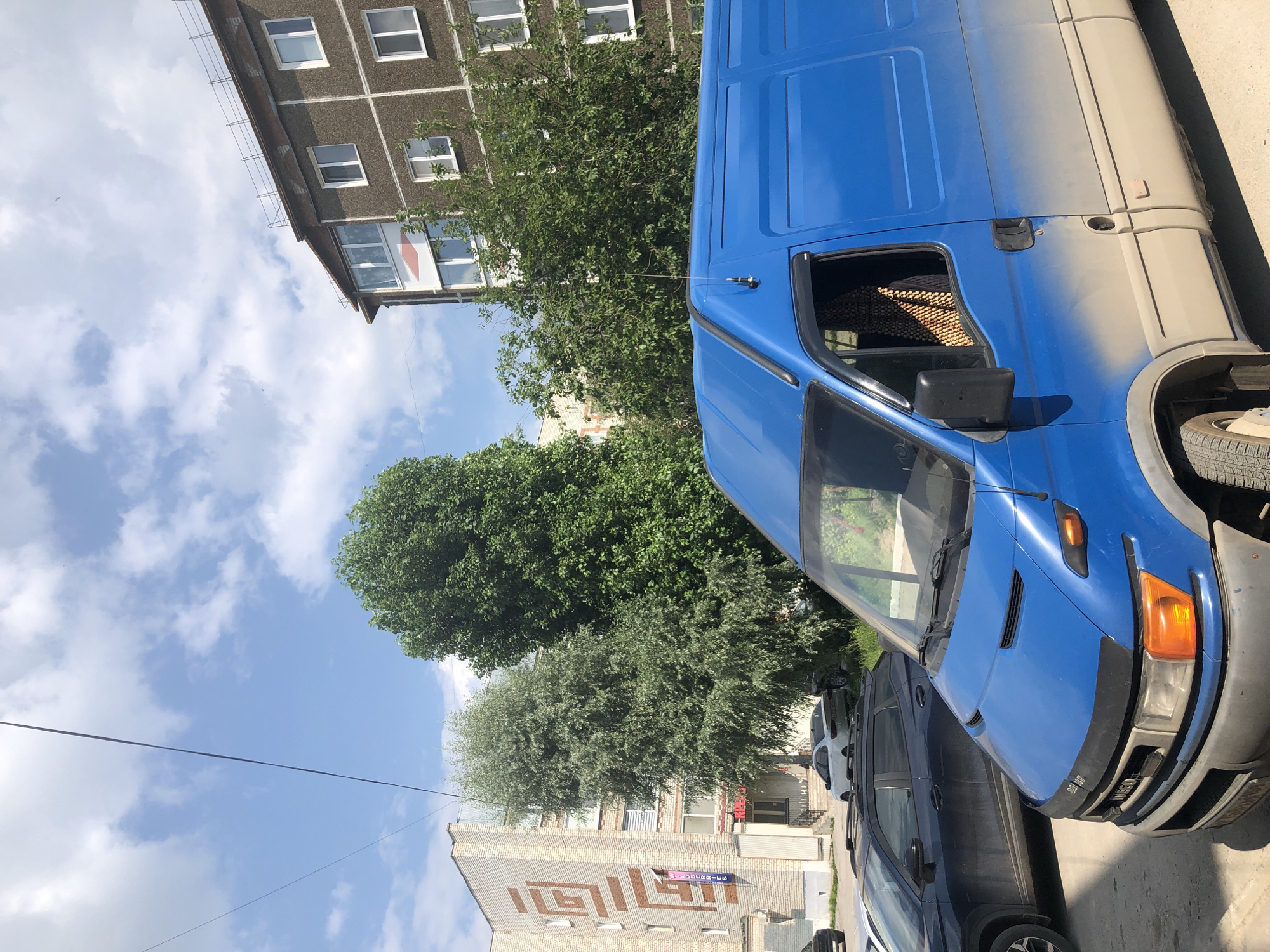 The image depicts a street scene in Solikamsk, Russia. In the foreground, a blue Iveco van is parked, showing significant dust and dirt accumulation on its lower half. The van has a black hood deflector. To the left of the van, a dark-colored SUV is parked, also appearing dusty. Behind the SUV, a white sedan is visible.
The background features two buildings. The building on the left is a multi-story brick structure with a geometric pattern on its facade. A blue sign with white lettering is visible on this building. The building on the right is a taller, multi-story apartment building with a grey, textured facade and several windows. Between the buildings, several large green trees are present, with one tree having silvery-green foliage and the other being densely leafed. The sky is blue with scattered white clouds, and power lines are visible traversing the upper portion of the image.