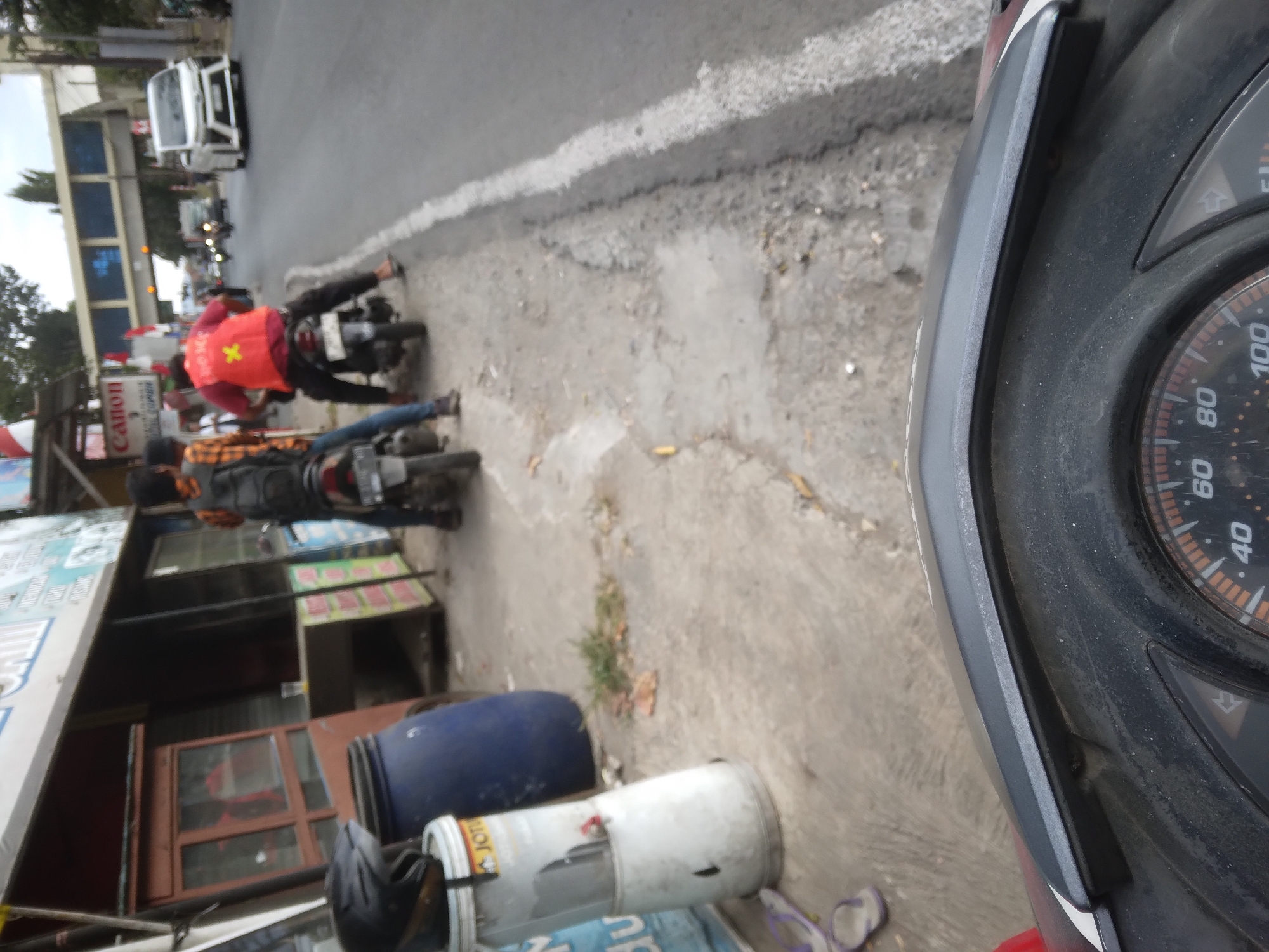 The image captures a scene on a street in Desa Pamijen, Kecamatan Sokaraja, Indonesia. In the foreground, the dashboard of a motorcycle is visible, with a speedometer displaying markings up to 100. The background shows two individuals on motorcycles, with one rider wearing a bright orange vest. To the left, a store with a red wooden cabinet and glass displays is situated along the roadside. A large blue barrel and a white cylindrical container are also present on the sidewalk. Further down the street, other vehicles, including a white van, are seen on the asphalt road, which is marked with a white line along the edge. An elevated pedestrian bridge is visible in the distance. The environment suggests an urban or semi-urban setting with elements of commerce and transportation.
