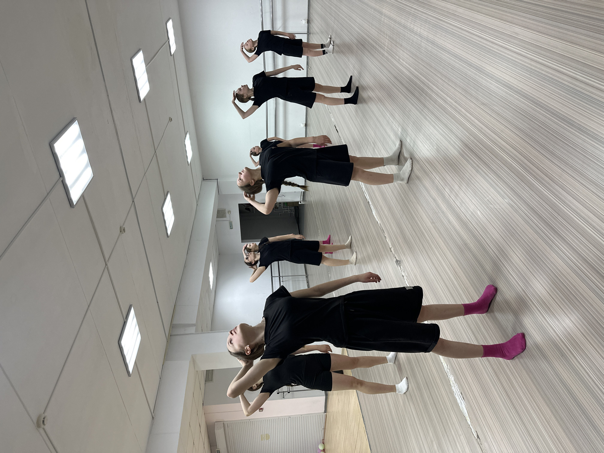 The image captures six young females engaged in a synchronized activity within what appears to be a dance studio or rehearsal space. The setting is indoors, brightly lit by multiple square fluorescent panels embedded in the ceiling. The floor is made of a light-colored wood or laminate with distinct horizontal striping.
The subjects, likely teenagers or young adults, are arranged in a line from the foreground to the background, all dressed uniformly in black short-sleeved tops and black shorts or skirts. Their footwear varies, with one participant sporting bright pink socks, others wearing white socks, and some with black socks or bare feet.
Each individual is performing the same pose: standing upright, with their heads tilted back, looking upwards, and their hands placed at the back of their heads or necks. This posture suggests a dance exercise, stretching, or a specific movement sequence. Their expressions are focused and serious, indicating concentration on the task. A ballet barre can be seen along the back wall, further reinforcing the studio environment. A large mirror covers the wall to the left, reflecting parts of the room and some of the participants.
On a distant wall, partially obscured, a white sign with red lettering reading "ВЫХОД" (Russian for "EXIT") is visible. The scene depicts a dedicated practice session, likely during the day or evening given the artificial lighting. The location context is Votkinsk, Russia.