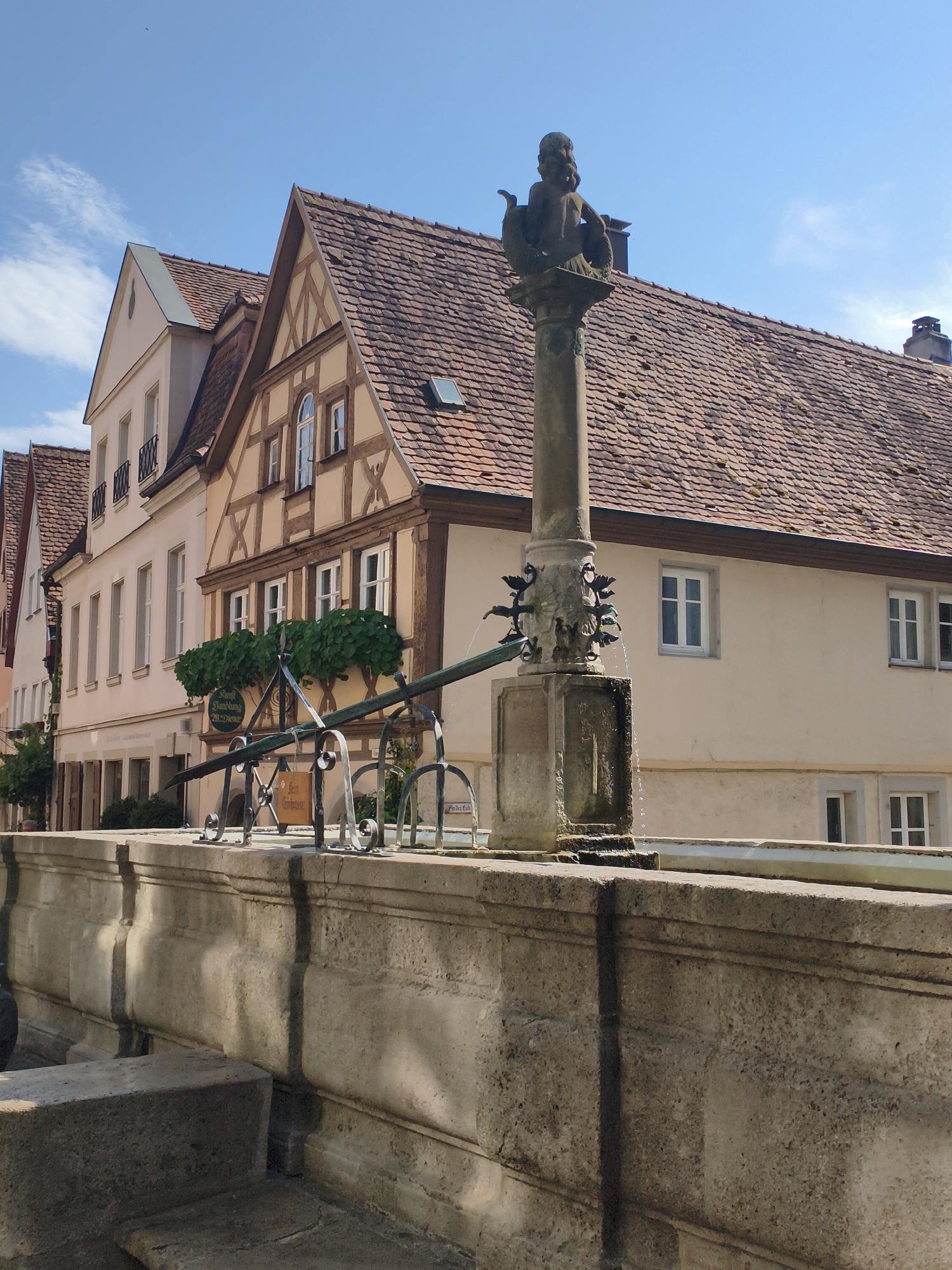 The image shows a view of a fountain in the historic German town of Rothenburg ob der Tauber. The fountain, made of stone, features a pillar with a statue of a mermaid-like figure at the top. Water spouts from several decorative gargoyles on the pillar. In the background, traditional half-timbered buildings with red-tiled roofs line a street. The sky is clear and blue with a few wispy clouds. The fountain appears to be active, with water flowing down its structure and into a basin. The overall scene depicts a picturesque streetscape characteristic of medieval German towns.
