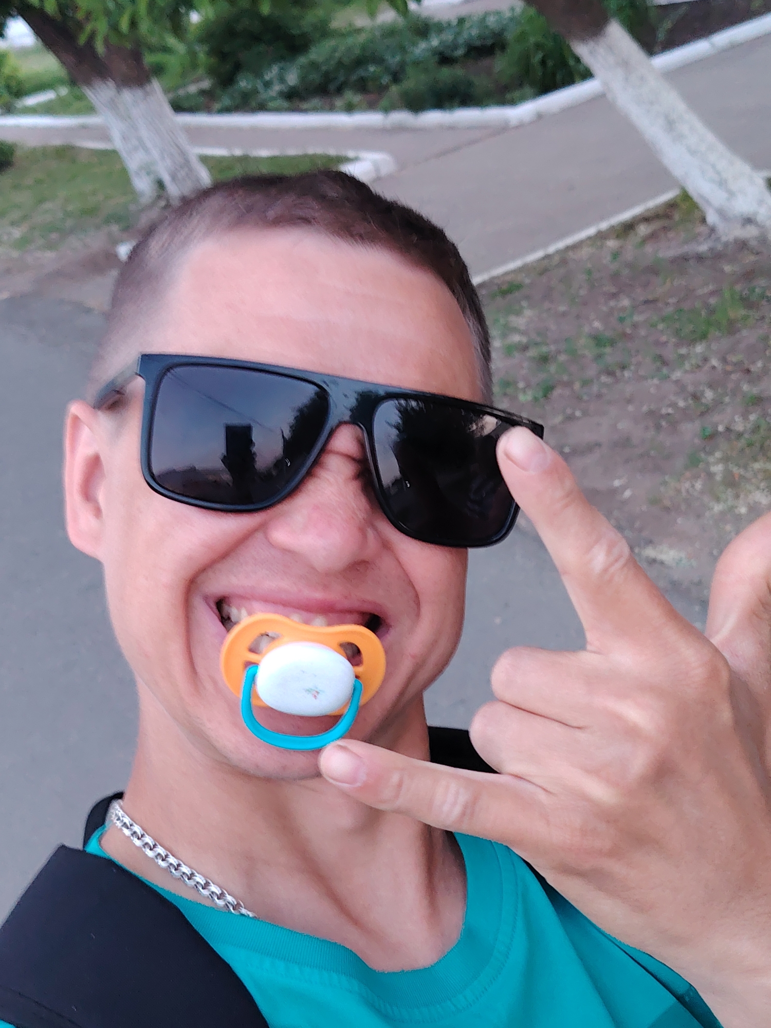 A selfie depicts a man outdoors, wearing black sunglasses and a teal shirt. He has short, dark hair and a pacifier in his mouth. His right hand is raised, with his index finger touching his sunglasses. A silver chain necklace is visible around his neck, and a black backpack strap is partially seen over his shoulder.
The background shows trees with whitewashed trunks and a gray sidewalk, suggesting an urban park or residential area in the city of Chaikovsky, Russia.