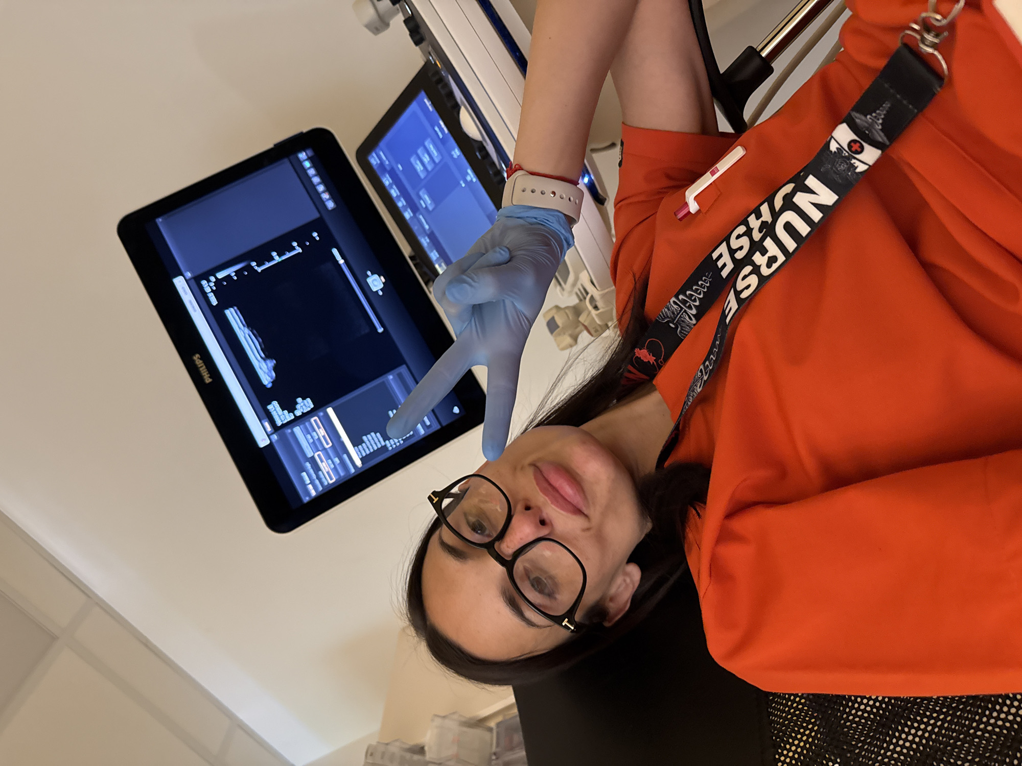 A young woman, likely a medical professional, is featured prominently in this image, taken in what appears to be a medical setting. She has dark hair and is wearing black-framed glasses and an orange scrub top. On her left wrist, she wears a white smartwatch or fitness tracker, and her right hand is covered by a light blue medical glove, forming a peace sign gesture. She looks towards the camera with a subtle smile.
Behind her, two medical monitors display complex medical imaging data, possibly from an ultrasound. The brand "Philips" is visible on the lower left screen. Various buttons and graphical elements typical of medical diagnostic equipment interfaces are discernible. Other parts of medical machinery are visible in the background. Around her neck hangs a black lanyard with the word "NURSE" printed multiple times in white, alongside medical-themed illustrations. A white pen is clipped to her scrub top. The overall setting is an indoor medical facility, such as a hospital or clinic.