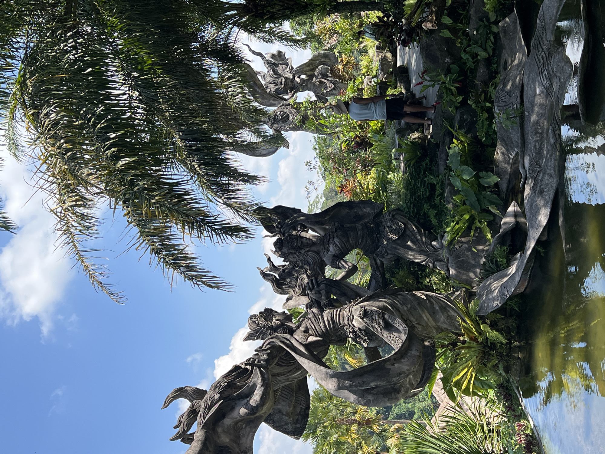 The image shows a daytime scene in Kedewatan, Indonesia. A weathered, gray stone sculpture depicting multiple figures stands prominently, partially reflected in the still water of a pond or waterway. Lush green vegetation surrounds the sculpture and pond. A palm tree with dense foliage is visible in the upper left of the frame, partially obscuring the blue sky dotted with white clouds. A person wearing a gray tank top and dark shorts stands on a walkway in the background.