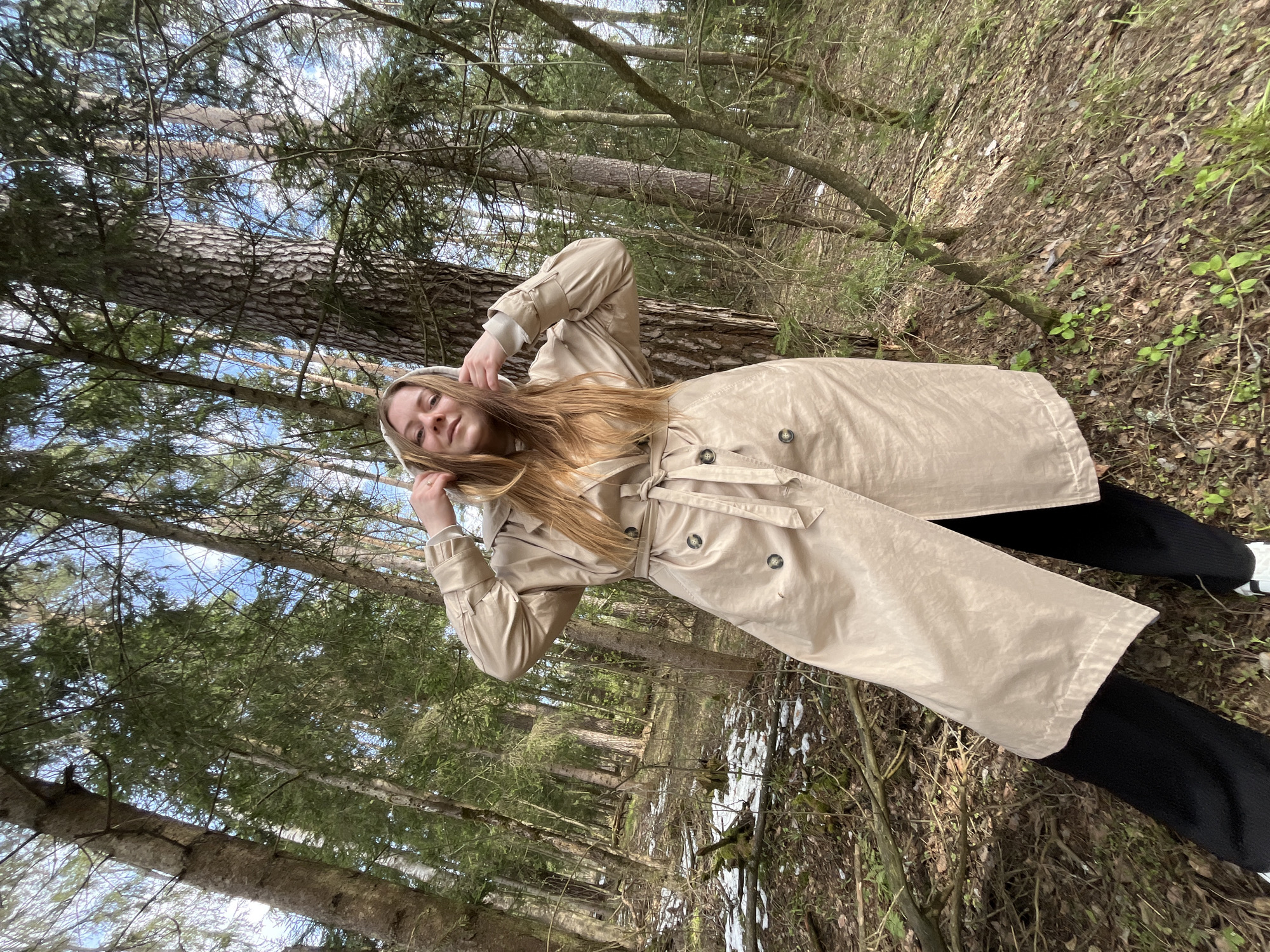 A young woman stands in a forest, wearing a beige trench coat over black pants. The location is likely in a wooded area near Раздолье, Russia, given the context. The woman has her hands near her head, possibly adjusting a hood or touching her hair. The environment consists of tall trees, some with bare branches, and ground covered with fallen leaves and patches of snow. The sky is visible through the branches.