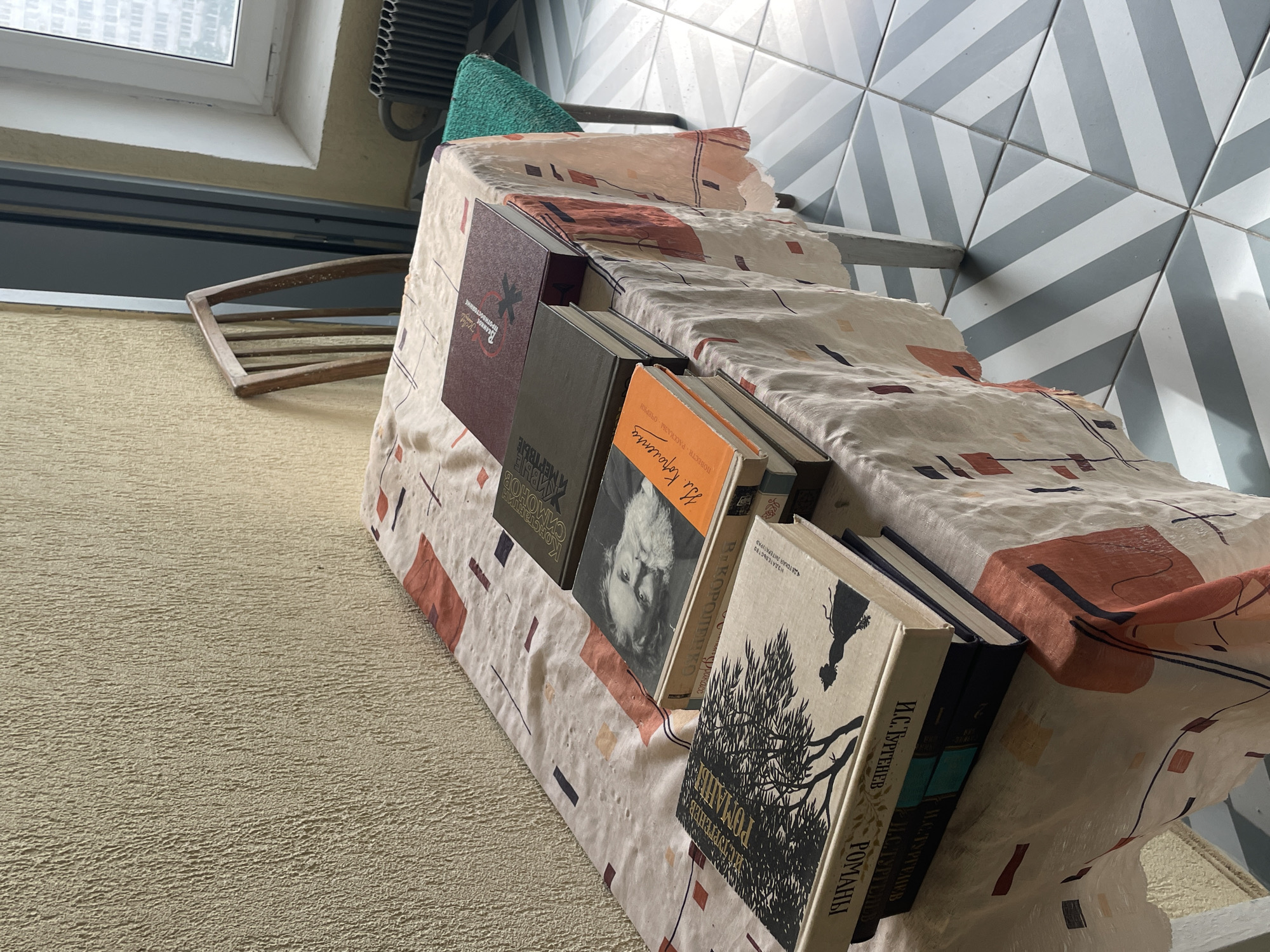 A close-up, eye-level shot shows a collection of books on a table covered with a light-colored tablecloth patterned with scattered squares and rectangles in reddish-orange, navy blue, and black. The books are stacked and arranged on the tablecloth. The leftmost book features a black silhouette of a tree and a figure against a light background on its cover. To its right is another book with a portrait of a man with a beard on its cover, and an orange spine with Russian text. Further to the right, another book has a dark gray cover and is topped by a burgundy book with Russian text and a red design. The table itself appears to be made of light-colored wood. Behind the table, a wooden chair with a tall, slatted back leans against a wall with a textured, light-colored finish. To the right of the chair, a window with white trim reveals a view of a city building, possibly in Moscow, Russia. A black radiator is visible below the window. The floor is tiled with a geometric pattern of light gray and white diagonal stripes. The lighting suggests it is daytime, with natural light entering from the window.