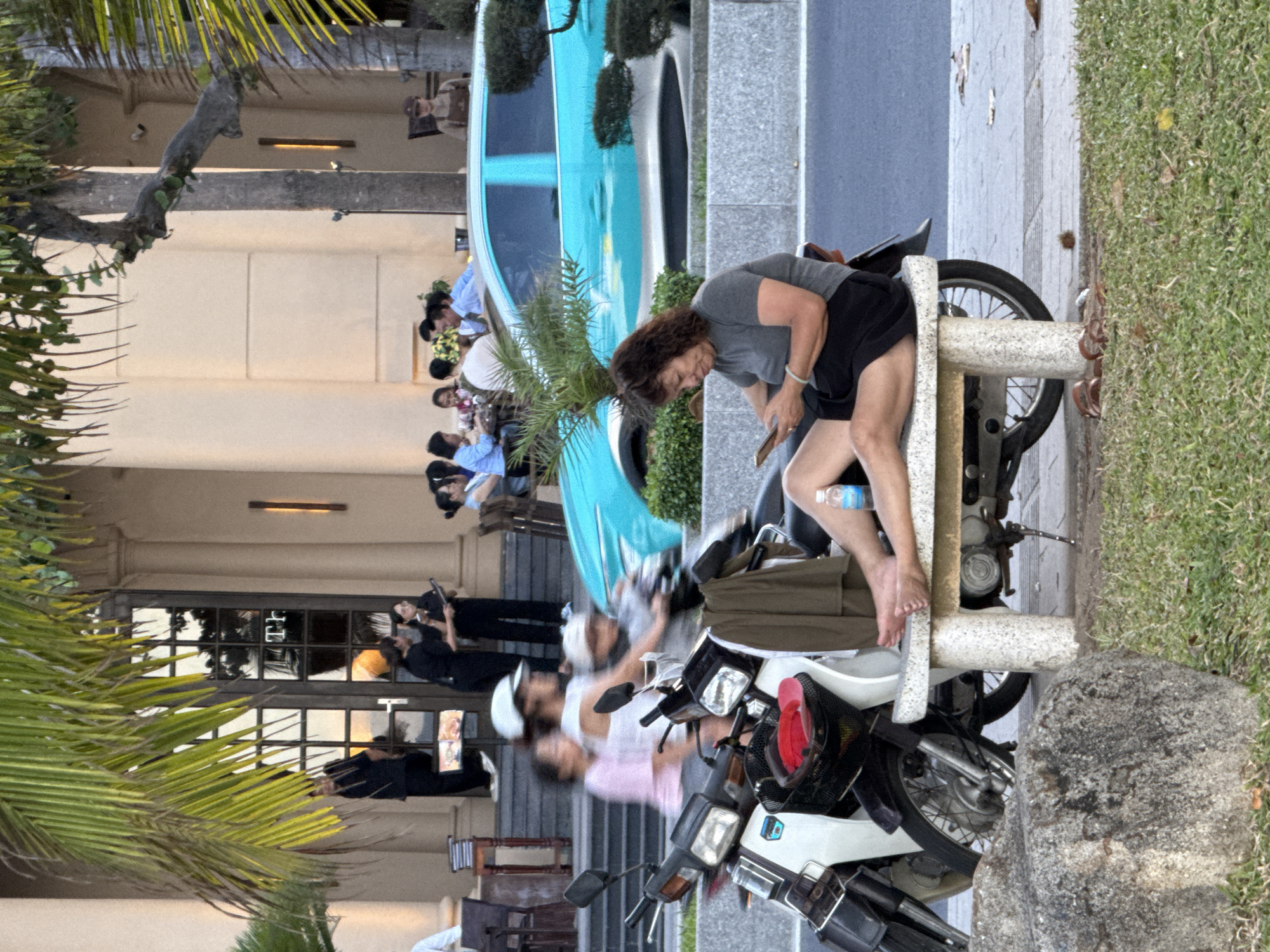 The image depicts a street scene in Nha Trang Ward, Vietnam, with a focus on individuals interacting with technology and their surroundings. In the foreground, a woman sits on a stone bench, engrossed in her mobile phone. Her feet are bare, and a bottle of water rests beside her. To her left, a white and black scooter is parked, with a green garment draped over its side. Behind the bench, a light blue car is visible, partially obscured by a palm tree.
In the middle ground, several people are gathered around tables outside a building, some appearing to be dining. Two servers in black uniforms stand near the entrance, one holding a tablet. Further back, individuals are seated at tables, engaged in conversation and enjoying their meals.
The background features a building with large glass doors and a signage that reads "TH." The architectural style suggests a commercial establishment, possibly a restaurant or cafe. Palm trees and other foliage are scattered throughout the scene, indicating a tropical or subtropical environment. The overall atmosphere is casual and lively, with a mix of individuals engaged in various activities.