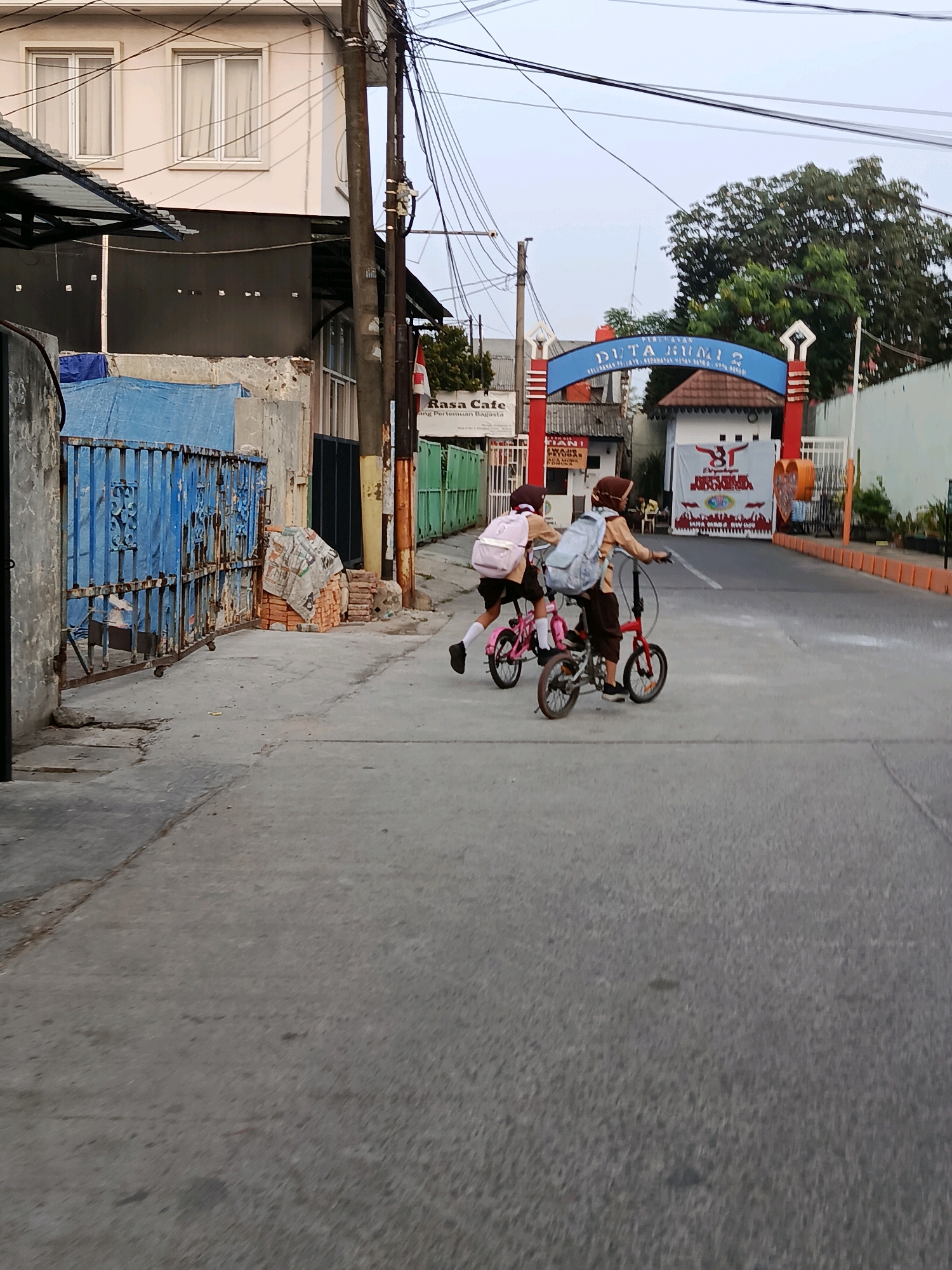 Two children wearing school uniforms and backpacks are riding bicycles on a paved road in Bekasi, Indonesia. The child on the left is riding a pink bicycle, and the child on the right is riding a red bicycle. Both children are wearing brown uniforms and headscarves. The road is lined with buildings and a fence on the left side, and a large archway with text reading "PERUMAHAN DUTA BUMI 2" on the right side. The sky is overcast. The overall scene depicts a typical residential street in Indonesia.