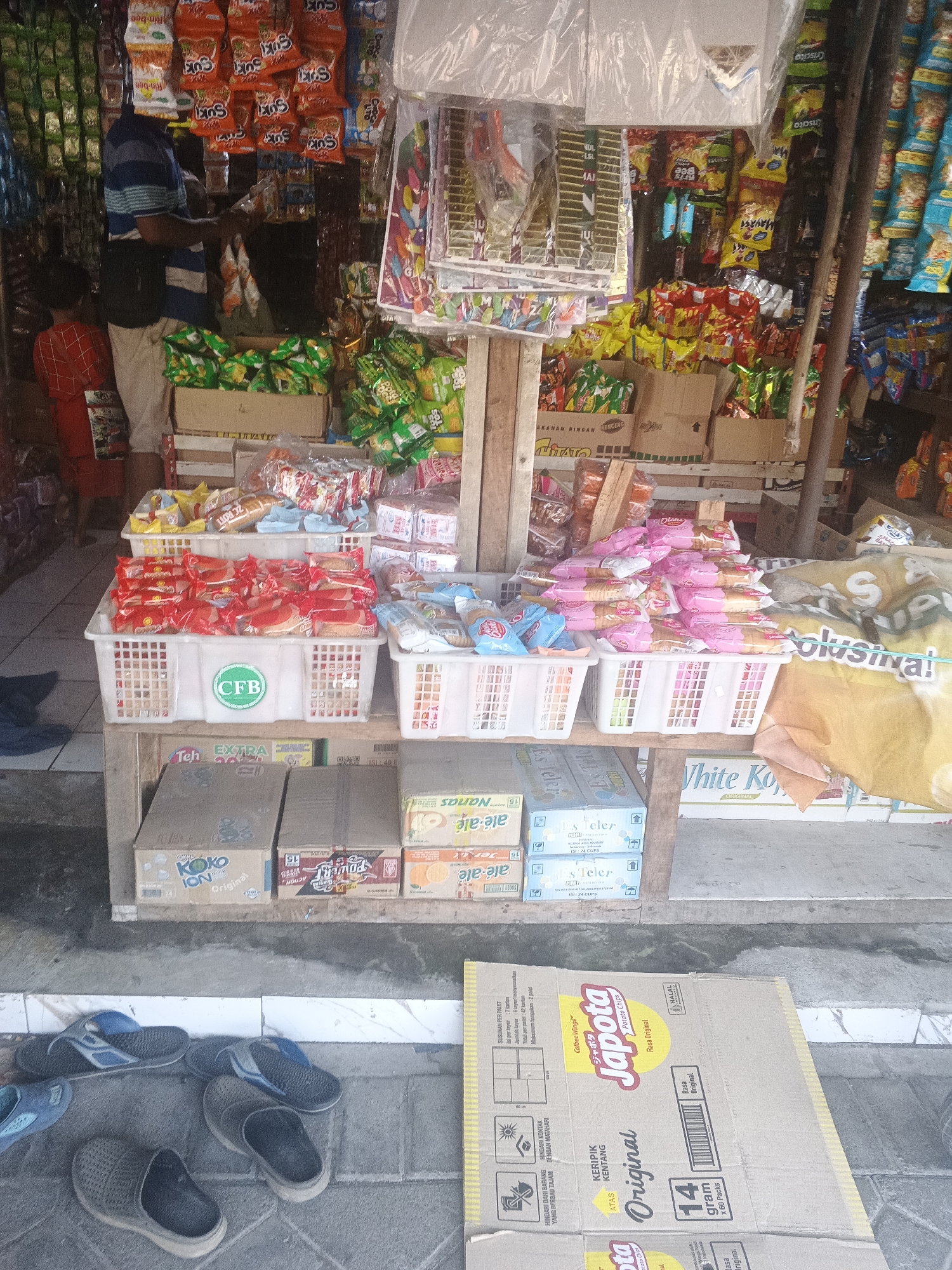 An outdoor, ground-level, medium shot captures a small, open-air convenience store in Krian, Indonesia, overflowing with packaged goods. A man wearing a blue and white striped shirt and beige pants stands behind a wooden stall, looking down at the merchandise. A child in a red shirt is partially visible on the left, facing away from the camera. The stall is packed with various snacks, chips, and drinks displayed in white plastic baskets and stacked cardboard boxes. Products include "CFB" branded items, "Koko Ion," "Teler," "Japota Potato Chips," and "White Kopi" coffee. The ground is paved with gray tiles. Several pairs of sandals and clogs are scattered on the ground in front of the stall. The overall scene depicts a busy, local retail environment.