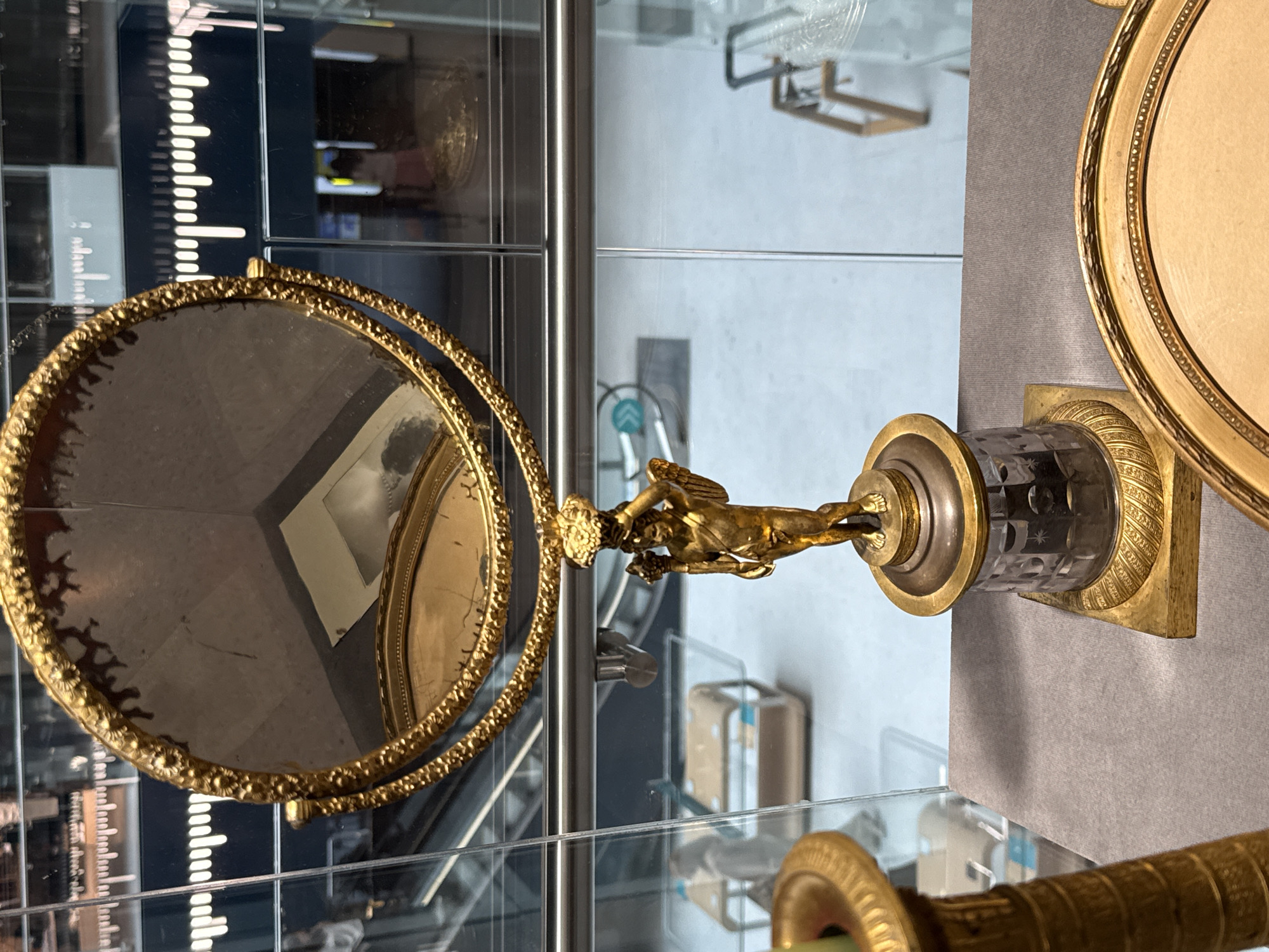 This media file displays a close-up, eye-level view of an ornate, gilded decorative object, likely a vanity mirror or a part of a larger display, situated within a museum or gallery setting. The object is predominantly gold, with intricate detailing on its frame and stand. The mirror itself is round and reflects a portion of the surrounding environment, including what appears to be another gilded decorative item and a display case with tiered shelving.
The central feature of the object is a golden statuette of a winged figure, possibly representing a mythological character like Cupid or a cherub, positioned above a cylindrical crystal element. This crystal element is faceted and appears to have an etched pattern. The statuette is posed dynamically, with one arm extended and the other bent, giving the impression of movement. The stand is supported by a rectangular base with decorative carvings.
In the background, through a glass partition, elements of the museum's interior are visible. There are shelves with various objects, some of which appear to be antique or artistic in nature. A distinctive feature in the background is a vertical scale or ruler with white markings against a dark background, possibly part of an exhibit or a measurement tool. The lighting suggests an indoor environment, likely illuminated by artificial lights. The overall impression is one of a preserved artifact presented for public viewing. No specific activity or event is taking place; the scene depicts a static object within a controlled exhibition space. No visible text is legible. The location context is Moscow, Russia.