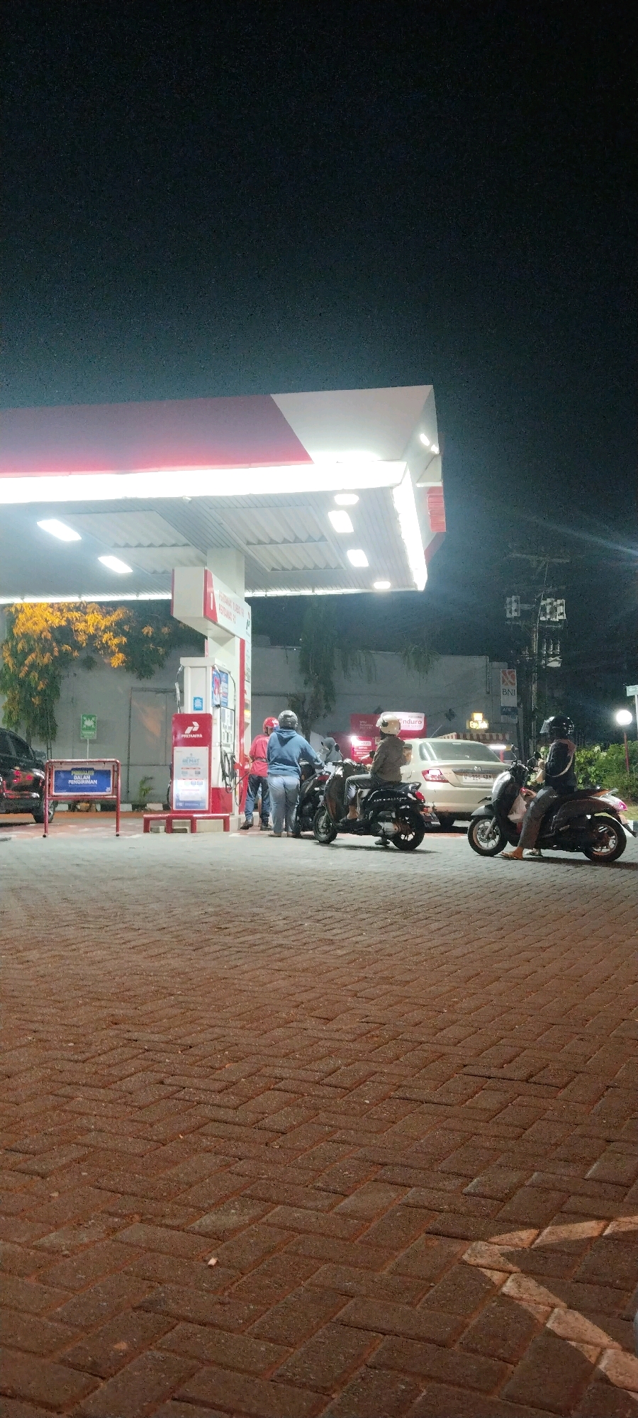 The image depicts a night scene at a Pertamina gas station in Karangturi, Indonesia. Several individuals are present, with three on motorcycles and two on foot near a gas pump. One person is on foot in a blue hoodie and jeans, standing beside a motorcycle. Another person in a red shirt is also on foot near the pump. A person wearing a white helmet and brown jacket is seated on a motorcycle to the right of the pump. Another individual in a black helmet and grey pants is also seated on a motorcycle to the right of the scene. A car is visible behind the motorcycles, and a building with a sign that reads "BNI" is in the background. The gas station has a red and white canopy illuminated by lights. The ground is paved with reddish-brown bricks. Trees with yellow foliage are visible to the left of the gas station.