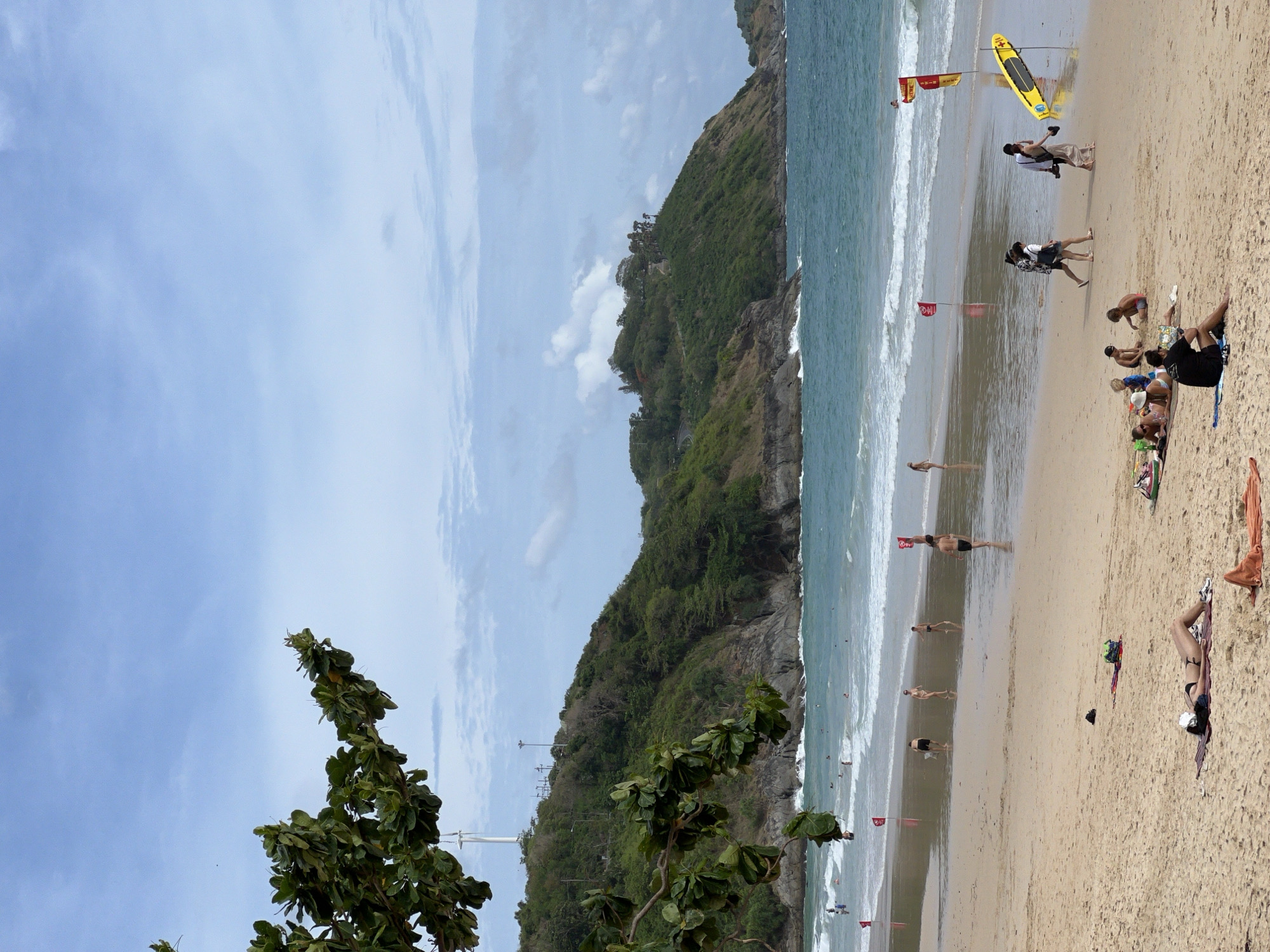 The image depicts a beach scene, likely in Unknown, Unknown, on a partly cloudy day. A green, tree-covered hillside borders the beach on the left side of the image. A sandy beach occupies the right side of the image, extending to the waterline. Numerous people are present on the beach, some sunbathing, others walking along the shore. Several red flags are placed in the sand near the water's edge. A yellow surfboard is positioned upright on the sand near the right edge of the frame.