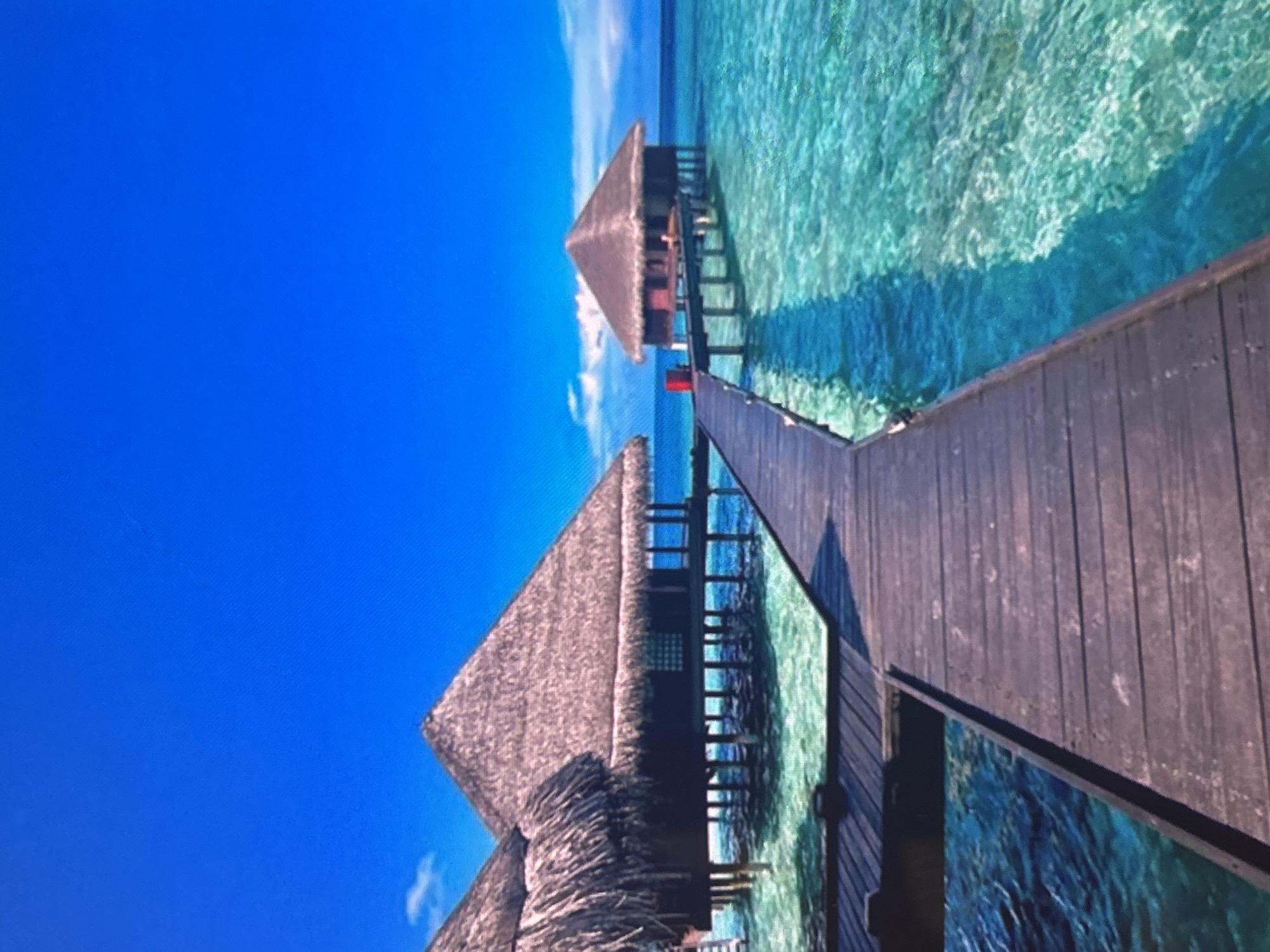 The image captures a vibrant tropical scene featuring overwater bungalows connected by a wooden pier, set under a brilliant blue sky. The picture appears to be rotated, but when viewed correctly, the horizon line separates the expansive blue sky from a clear, turquoise body of water, likely an ocean or lagoon.
Two overwater bungalows with traditional thatched roofs are prominent. One is closer to the foreground on the left, partially visible, while another is situated further along the pier. Both structures are elevated on stilts above the water. A dark wooden pier or walkway extends diagonally from the lower right of the frame towards the background, linking the bungalows.
The water below and around the structures is exceptionally clear, exhibiting various shades of blue and green, with sunlight reflecting on its surface and revealing patterns on the seabed. Shadows from the bungalows and pier are cast onto the water, indicating bright, direct sunlight. The sky is a deep, unblemished blue with only a few faint, wispy clouds near the horizon.
No people are visible in the scene, and no specific activity is taking place, creating a tranquil and idyllic atmosphere. The setting suggests a remote, luxurious tropical resort, likely during daytime under sunny weather. There is no discernible text present in the image.
