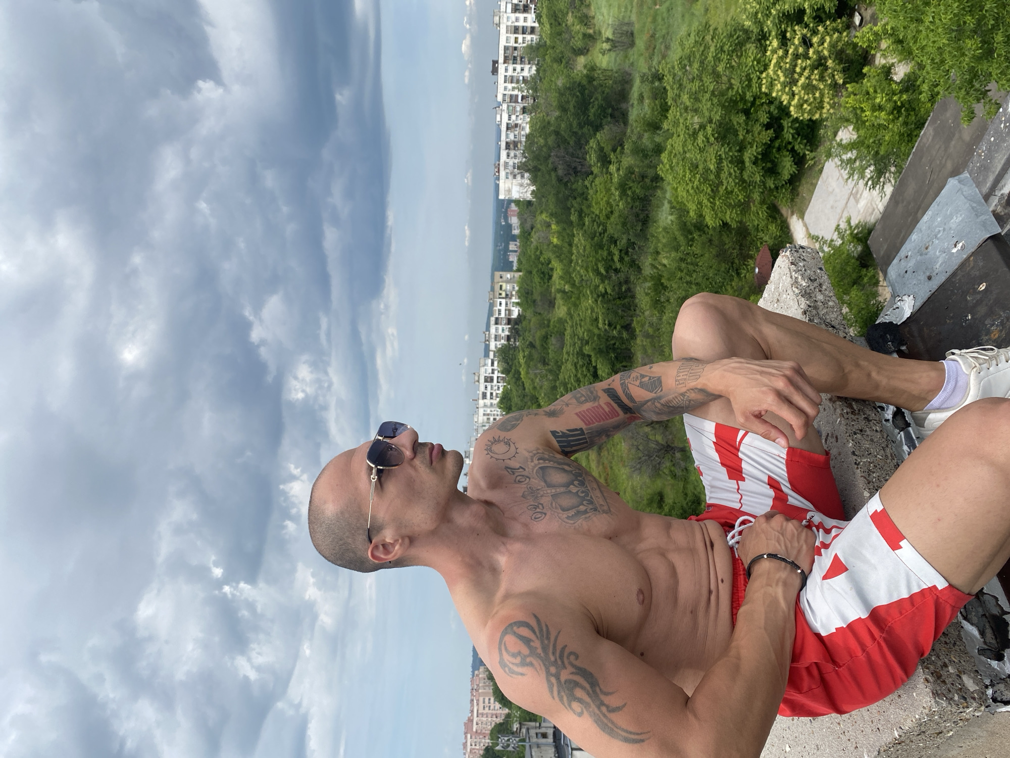 A man with a shaved head and sunglasses is sitting on the edge of a rooftop overlooking a city. He is shirtless and wearing red and white shorts. The man has tattoos on his arms and chest. In the background, there are buildings and trees. The sky is cloudy. The location is the city of Pleven, Bulgaria.
