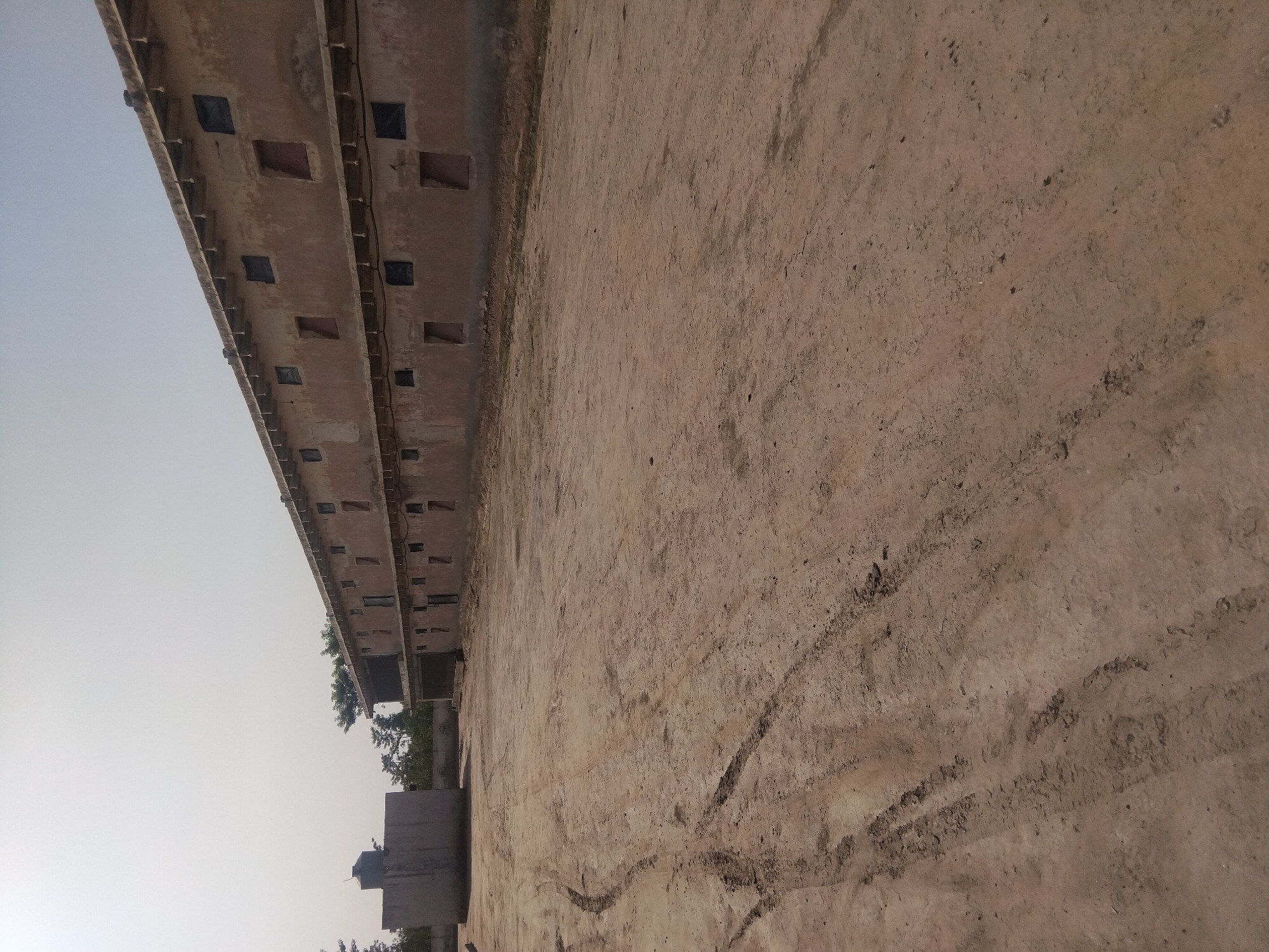 The image shows an exterior view of a long, two-story building under construction in Haveli Jhangranwali, Pakistan. The building's facade is a light brown, unplastered brick, with dark-covered openings that appear to be windows and doorways. Small, triangular roof supports jut out from beneath the upper level's overhang. A large, concrete water tank or silo stands to the left of the building, and further back, trees and a fence are visible. The ground in front of the building is a dusty, dirt lot with visible tire tracks. The sky is a pale, hazy blue.