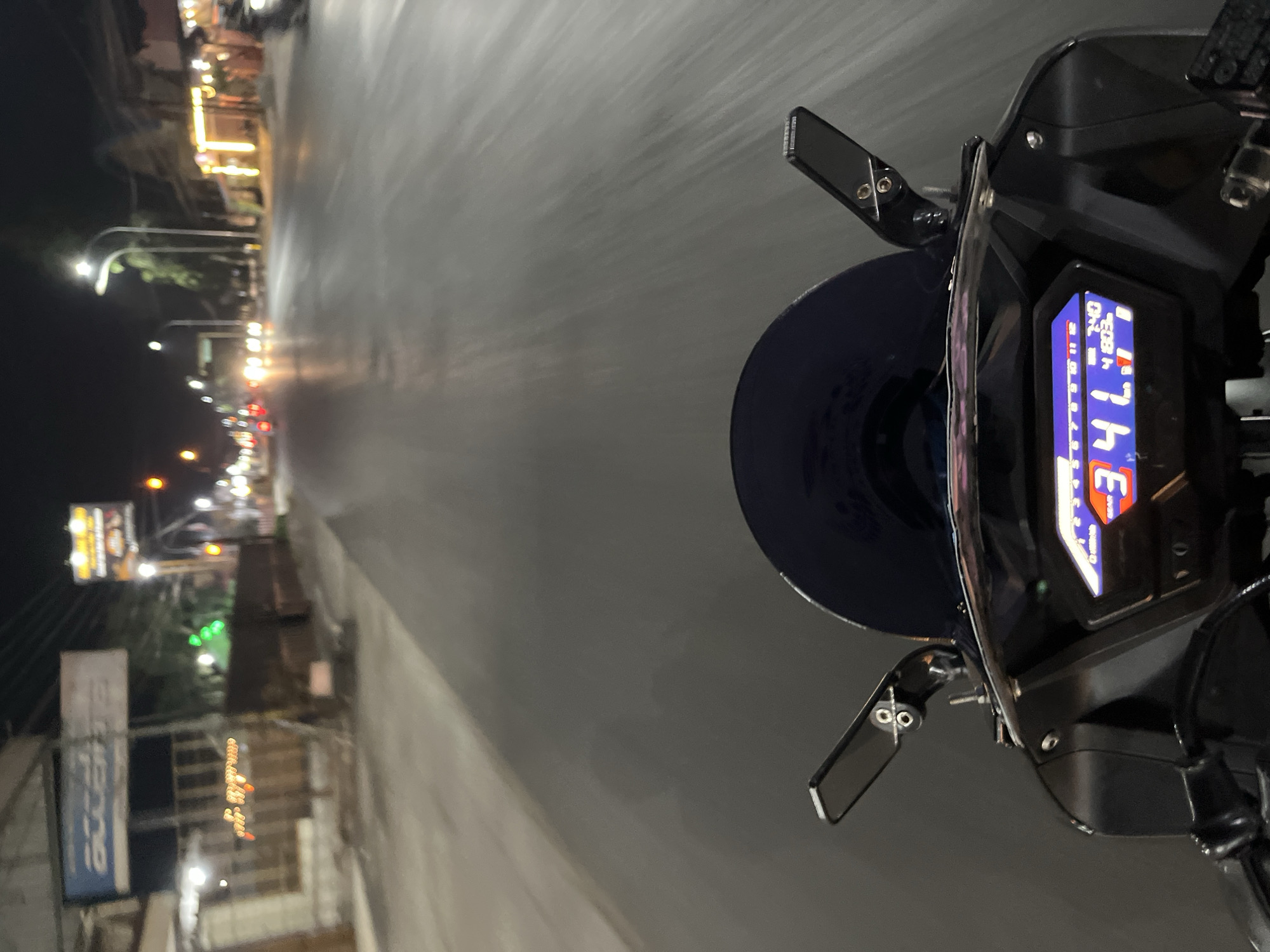 The image is a first-person perspective from a motorcycle at night on a street in Rejosari, Indonesia. The motorcycle's digital dashboard is visible in the foreground, displaying "34" as the speed in km/h, a gear indicator of "3", and a mileage reading of "41803". The road ahead is wet and reflects the lights of passing vehicles and streetlights. Buildings line both sides of the street, with some illuminated by yellow lights, suggesting businesses or residences. A blue windscreen is also visible above the dashboard. The overall scene depicts nighttime urban traffic in a Southeast Asian setting.