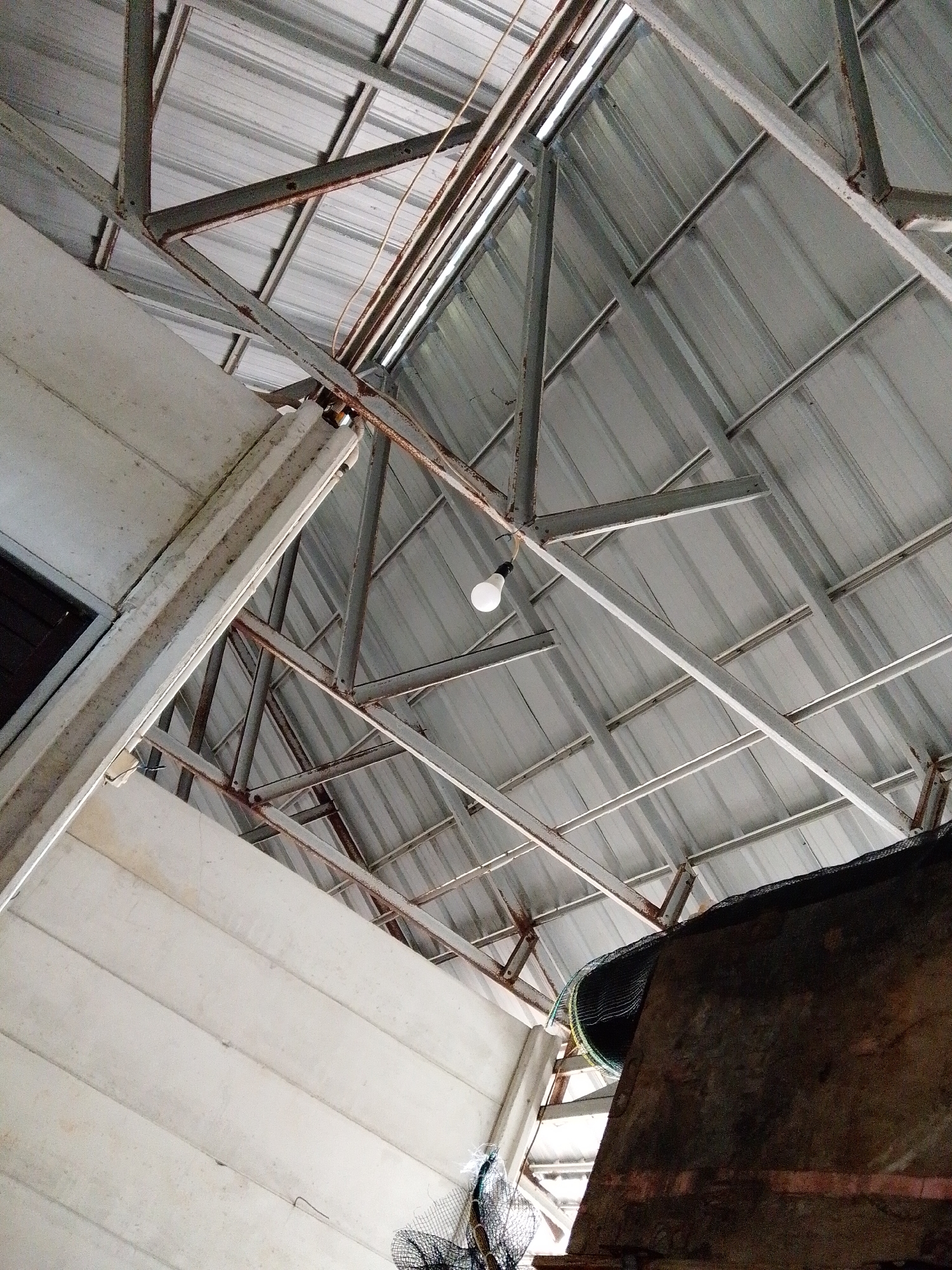 The image shows the underside of a corrugated metal roof supported by a metal framework. A single lightbulb hangs from one of the metal beams. Below the roof, a section of a white wall is visible. The bottom right corner of the image features a dark, possibly metallic, structure covered with a black mesh. The location is Bukit Ketok, Indonesia. No people are visible.