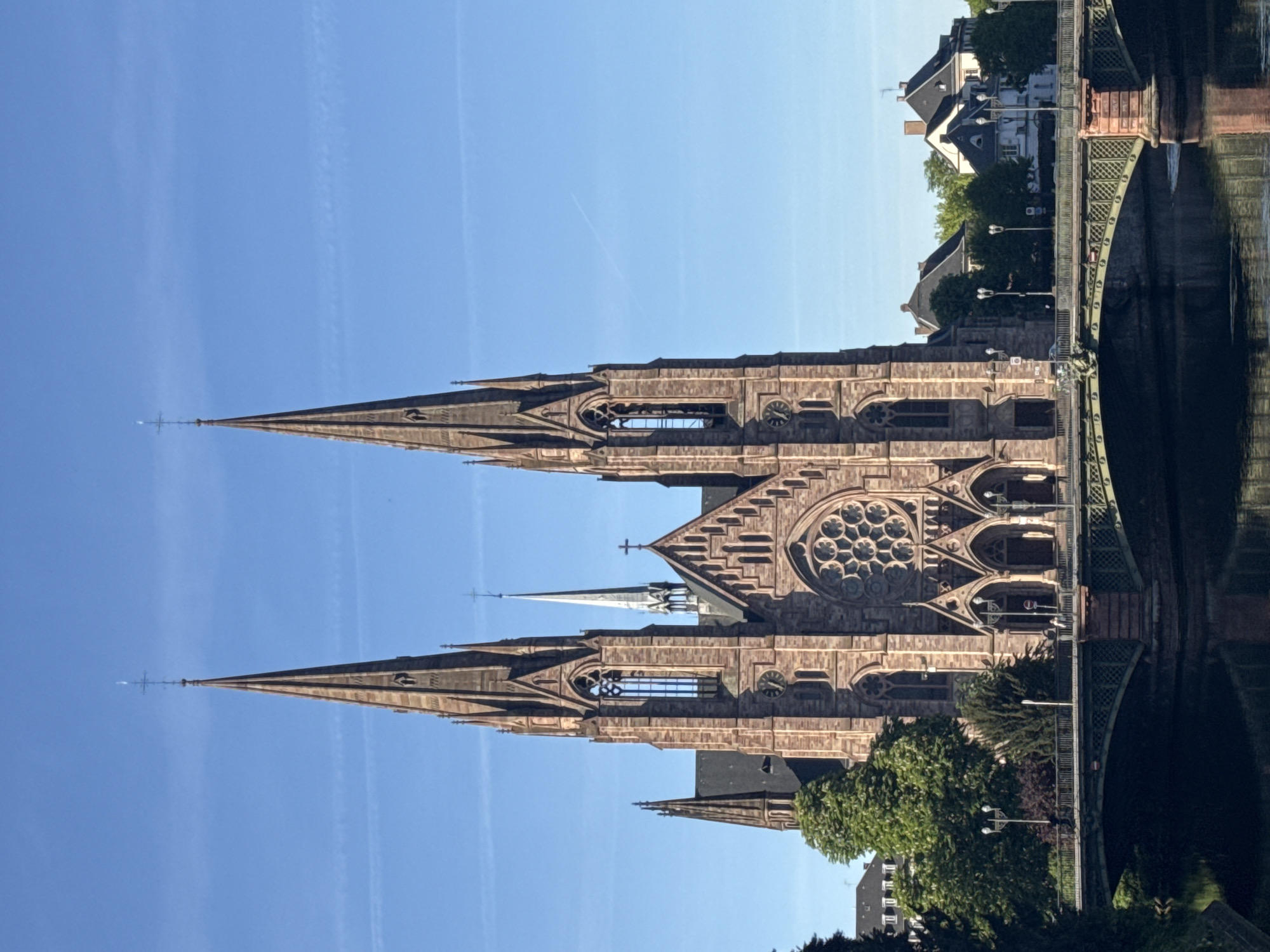 An imposing Gothic Revival church dominates the scene, featuring two tall, pointed spires made of reddish-brown stone. Each spire is adorned with intricate architectural details, including lancet windows and decorative elements, and is topped with a cross. A large rose window is visible on the church's facade, along with other arched windows. The church stands beside a body of water, likely a river or canal, in which its grand structure is clearly reflected, indicating calm conditions.
To the right of the church, a decorative metal bridge with green-painted supports and arches spans the water. Beyond the bridge, several traditional European buildings with varied rooflines are visible, nestled among green trees and lush vegetation lining the waterway. The sky above is a clear, bright blue with a few faint white contrails, suggesting excellent weather. The strong sunlight indicates it is daytime, likely late morning or early afternoon, casting distinct shadows on the church and surrounding elements.
No specific activity or people are prominent, giving the scene a tranquil and timeless quality. The architecture strongly suggests a historical European city. A visible red circular sign on the bridge with a white horizontal bar indicates a "No Entry" or "Do Not Enter" restriction, possibly for vehicles. Given the architectural style and riverine setting, this scene is consistent with the historic city of Strasbourg, France.
