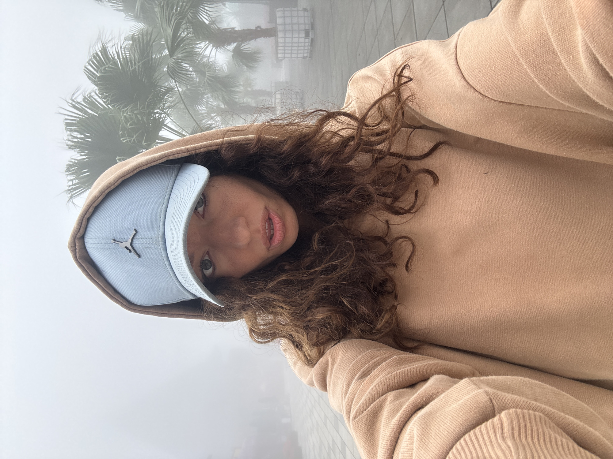 A young woman is taking a selfie in Đà Nẵng, Vietnam. She is wearing a tan hoodie and a light blue baseball cap with a Jordan logo. She has curly brown hair. The background is foggy, with palm trees and indistinct buildings visible.
