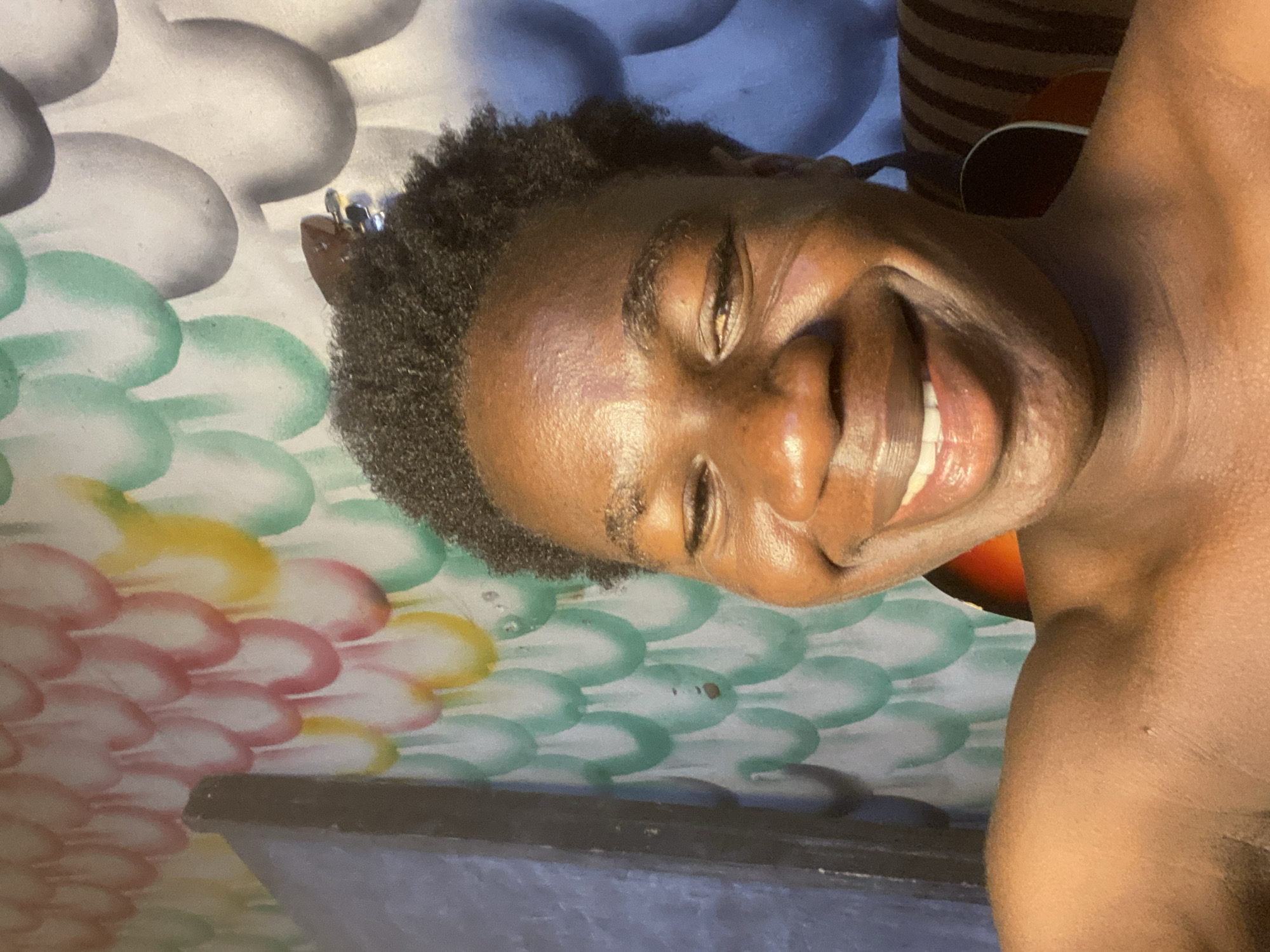 A smiling dark-skinned person with short hair is lying on a surface with a colorful, cloud-like pattern. The colors of the pattern include gray, blue, green, yellow, and pink. The person is smiling broadly, exposing their teeth. A portion of their shoulder is visible. An object, possibly a guitar, is partially visible in the background. The setting is indeterminate, but it is stated to be Mararaba, Nigeria.