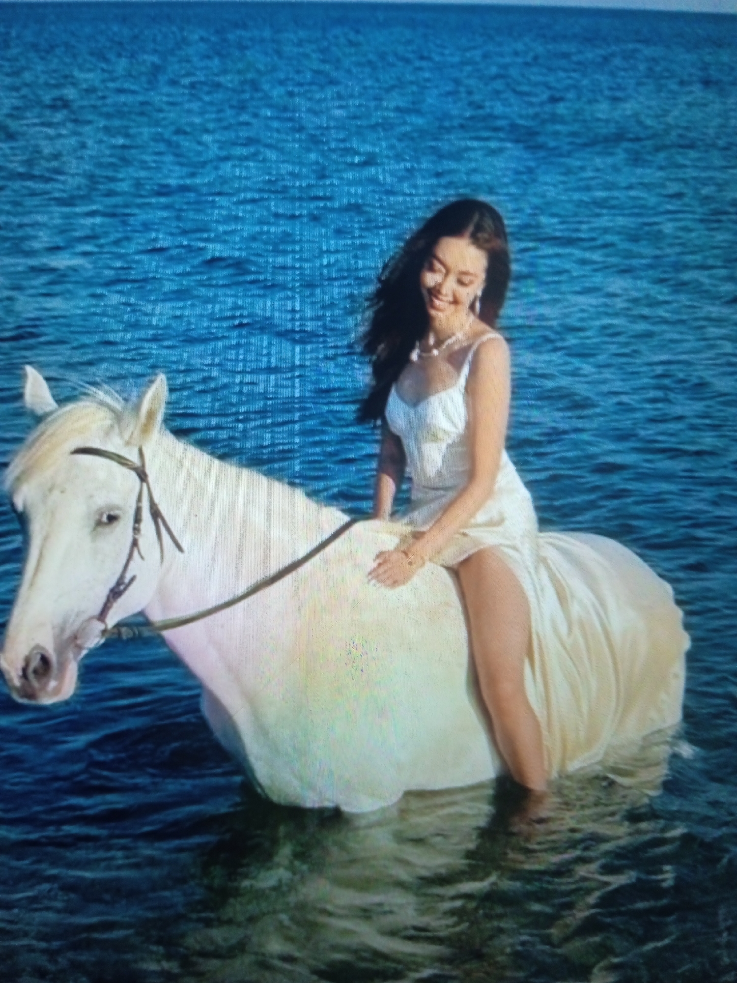 A young woman with long dark hair, wearing a white slip dress and a pearl necklace, is riding a white horse through shallow blue water. The horse's head is turned to the left, with its ears perked. The woman is smiling and looking down. The water is calm, with small ripples. The background is a vast expanse of blue water. The scene is serene and ethereal, evoking a sense of fantasy or dream. The exact location in Chernivtsi, Ukraine, is not discernible from the image.