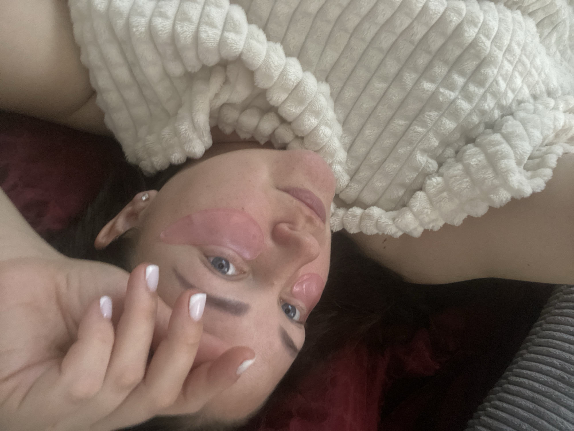 A woman with pink eye patches lies on a maroon surface, partially covered by a white, corduroy blanket. Her hand is raised near her face, displaying nails with white tips. The location is not discernible from the image.