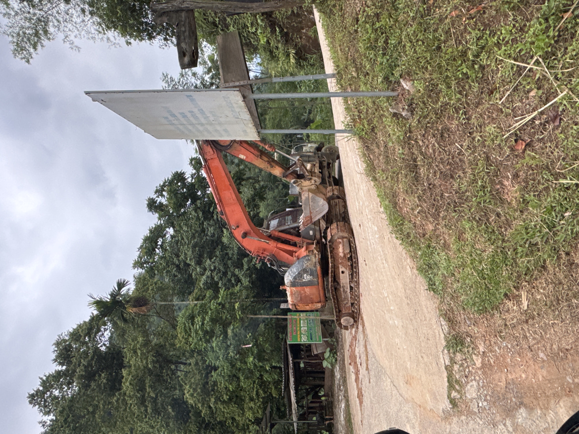 The image depicts an excavator positioned on a dirt and concrete road in Cổ Lũng Commune, Vietnam. The excavator, a large orange machine with a metallic bucket, is stationary and appears to be in a working or parked state. To the left of the excavator, a small, rustic-looking structure with a corrugated metal roof is visible, suggesting a rural or agricultural setting. A green sign with text and illustrations stands near the excavator, indicating information or instructions. A tall, white sign with some text is positioned to the right of the excavator, further down the road. The environment is characterized by lush green trees and vegetation covering hillsides in the background, under an overcast sky. The ground consists of a mixture of soil and a paved or concrete surface. There are no people clearly visible in the image. The scene conveys a sense of construction or ongoing work in a natural, mountainous landscape.