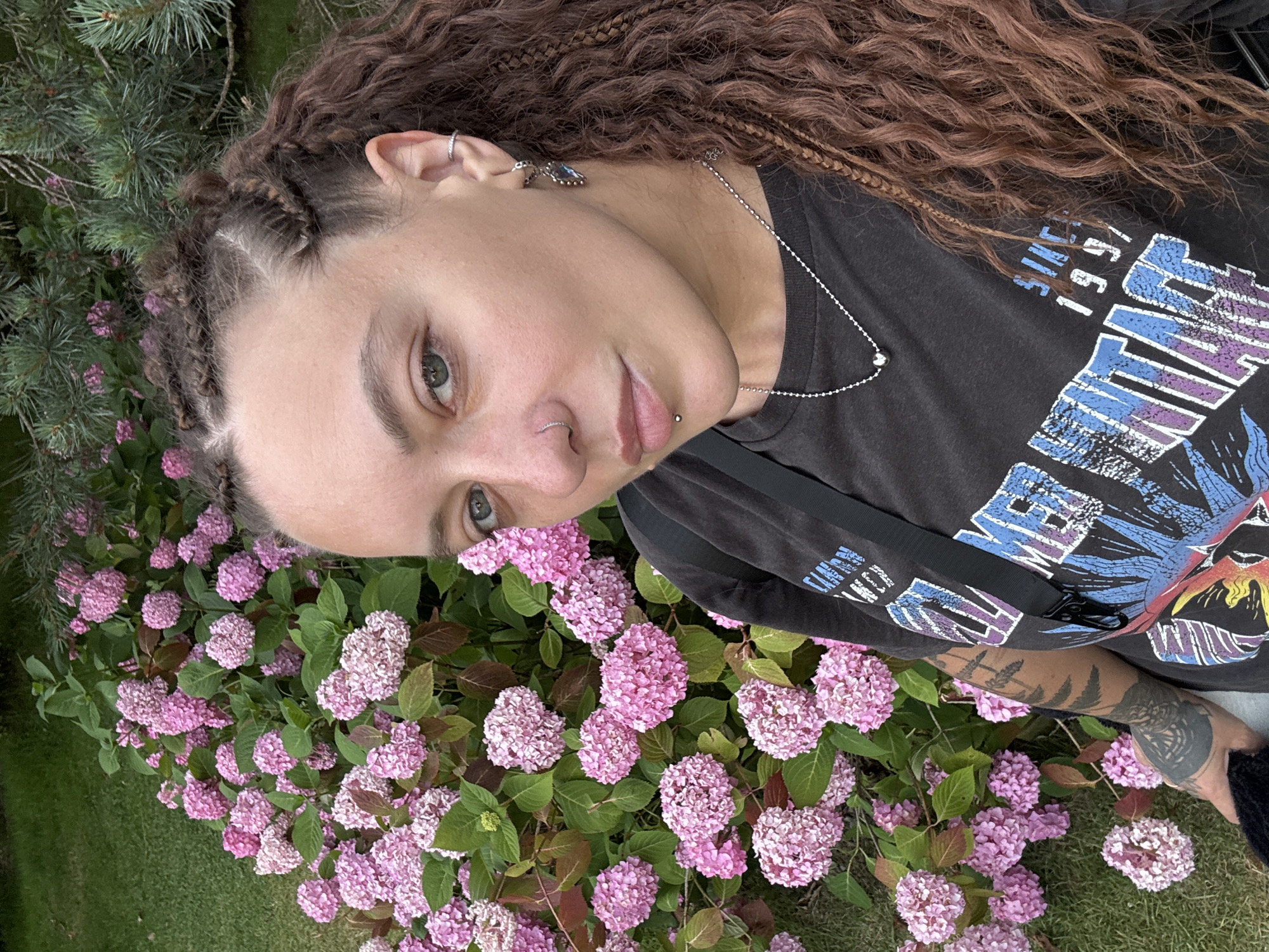In a medium-shot selfie, a person with long, curly brown hair, styled with braids near the forehead, looks towards the camera. They have a nose ring and a lip piercing. The person is wearing a dark t-shirt with a graphic print and a silver necklace. Their left arm, visible on the left side of the frame, is tattooed. Behind the person is a dense cluster of pink hydrangeas in full bloom, with green foliage. A pine tree with blue-green needles is visible in the upper right background. The setting appears to be an outdoor garden, likely in the city of Sochi, Russia, given the context.