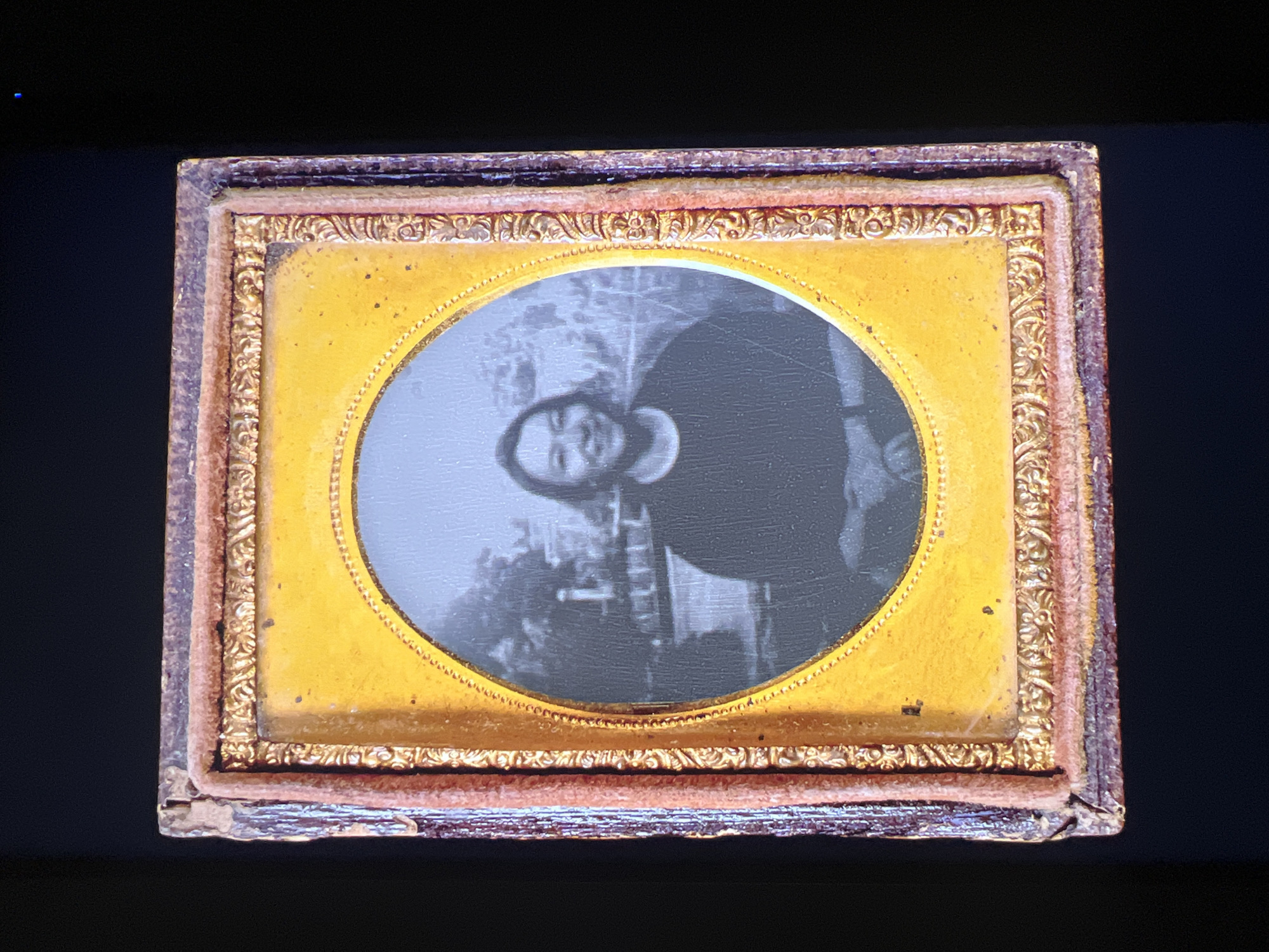 This media file presents an antique oval-shaped photographic portrait, possibly an ambrotype or tintype, encased within an ornate, rectangular frame. The artifact is displayed against a solid black background.
The frame consists of an outer structure, appearing to be dark wood or a similar material, with a reddish-brown, velvet-like inner lining that shows signs of wear and age, especially at the corners. Inside this, a gilded metal mat with intricate scrollwork patterns frames the photograph. The mat also features a smooth, bright gold band and a delicate beaded border immediately surrounding the oval image.
The photograph itself is a black-and-white or silvery-grey portrait of a single individual, likely a woman, captured from the head to the shoulders. She gazes directly at the viewer with a neutral, composed expression, characteristic of 19th-century portraiture. Her dark hair is styled, and she wears a simple, dark-colored, high-necked garment. The background within the photographic image is softly blurred, showing indistinct details that suggest an outdoor setting or a painted backdrop, with shapes resembling foliage or trees on the left and a building structure on the right.
The overall appearance of the item suggests it dates from the mid-to-late 19th century, indicating a formal portrait sitting. The frame's condition highlights its age and historical value. No visible text is present on the photograph or its frame. This historical artifact is located in Saint Petersburg, Russia.