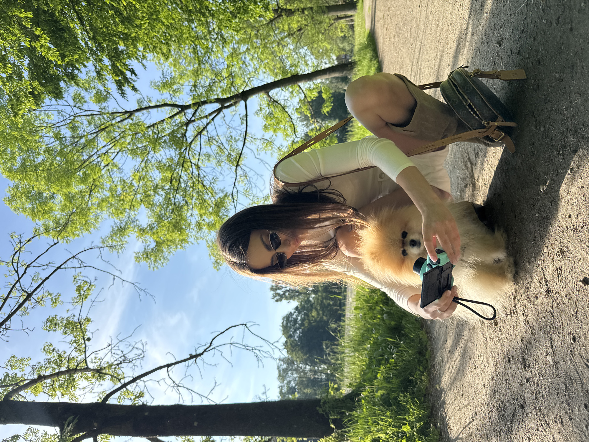 A woman is squatting on a dirt path, holding a small digital camera and aiming it at a Pomeranian dog. She is wearing sunglasses, a light-colored long-sleeved shirt, and dark shorts. A purse rests on the ground next to her. The scene is outdoors, with trees and sunlight visible in the background. The location appears to be a park path in Unknown, Unknown.