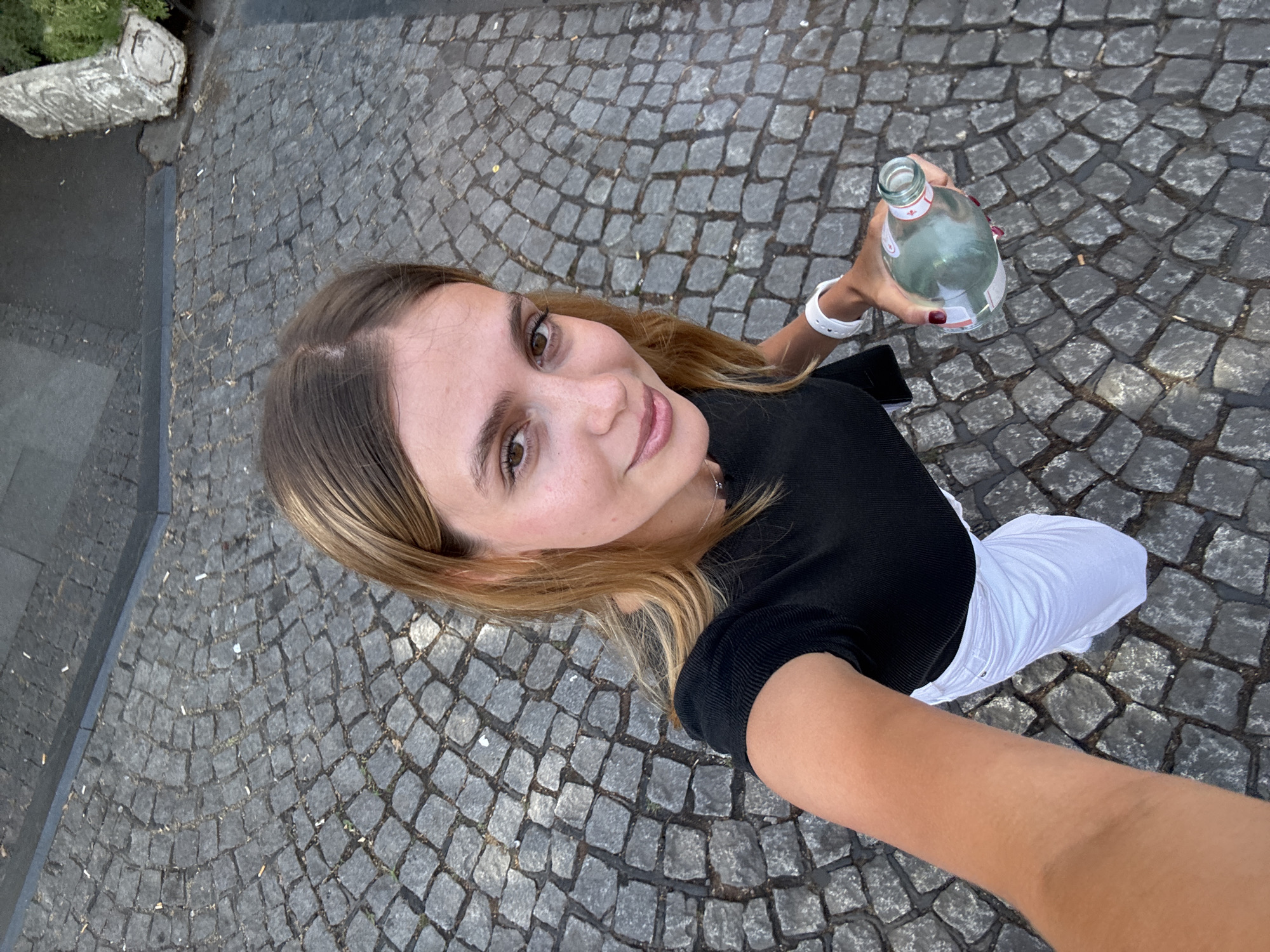 A young woman with blonde hair is taking a selfie outdoors on a cobblestone street. She is wearing a black short-sleeved top and white pants. She is holding a clear glass bottle of water in her right hand and a white smartwatch on her left wrist. The background is a textured cobblestone pavement. There are scattered cigarette butts on the ground. A concrete structure and some greenery are visible in the upper right corner. The scene appears to be in Belgrade, Serbia.