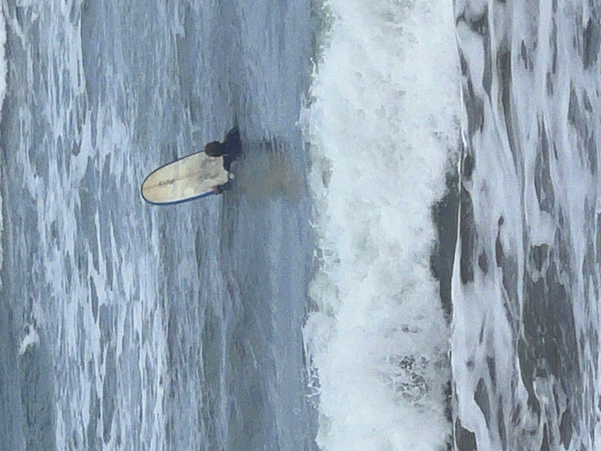 An image, originally oriented vertically but depicting a horizontal scene, captures a surfer on a light-colored surfboard in the ocean near Las Palmas De Gran Canaria, Spain. The surfer, visible from an elevated or overhead perspective, is dressed in a dark wetsuit or rashguard, with only their head and upper back visible as they lie prone on the board. The surfboard features blue rails and what appears to be faint, illegible text on its deck. To the right of the surfer, a large, foamy white wave is actively breaking, dominating a significant portion of the frame with its powerful churn. The water to the left of the surfer is a calmer, textured blue-gray, showing the movement of the sea. The scene takes place during the daytime, under what appears to be an overcast sky, resulting in soft, diffused lighting across the water. The image focuses on the dynamic interaction between the surfer and the ocean's waves.