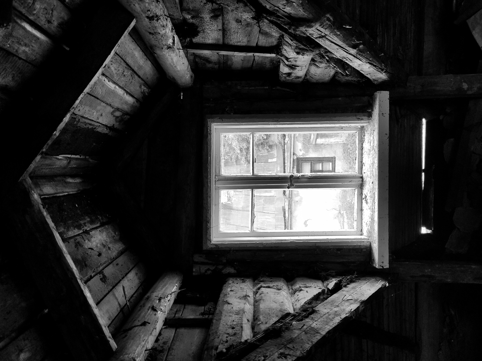 The image shows the interior of a dilapidated attic in black and white. The room is constructed primarily of rough, unfinished wood. A window is present, showing an external view that suggests an urban environment, possibly in Hrodna, Belarus. The exterior view features a building across from the window. The attic appears to be in a state of disrepair, with visible gaps in the wooden structure and debris scattered on the floor. The overall impression is one of neglect and decay.