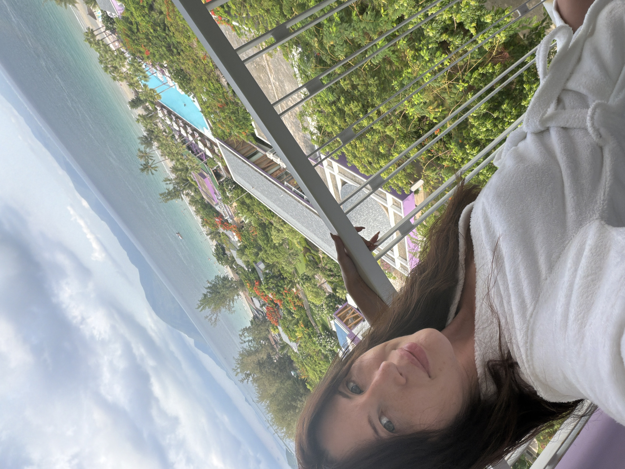 A woman with long, dark hair is taking a selfie on a balcony overlooking a resort. She is wearing a white bathrobe. The resort features a swimming pool, palm trees, and lush green vegetation with red flowers. Beyond the resort, a beach and the ocean are visible, with mountains in the distance. The sky is cloudy. The location is identified as Dong Ninh Hoa Ward, Vietnam.
