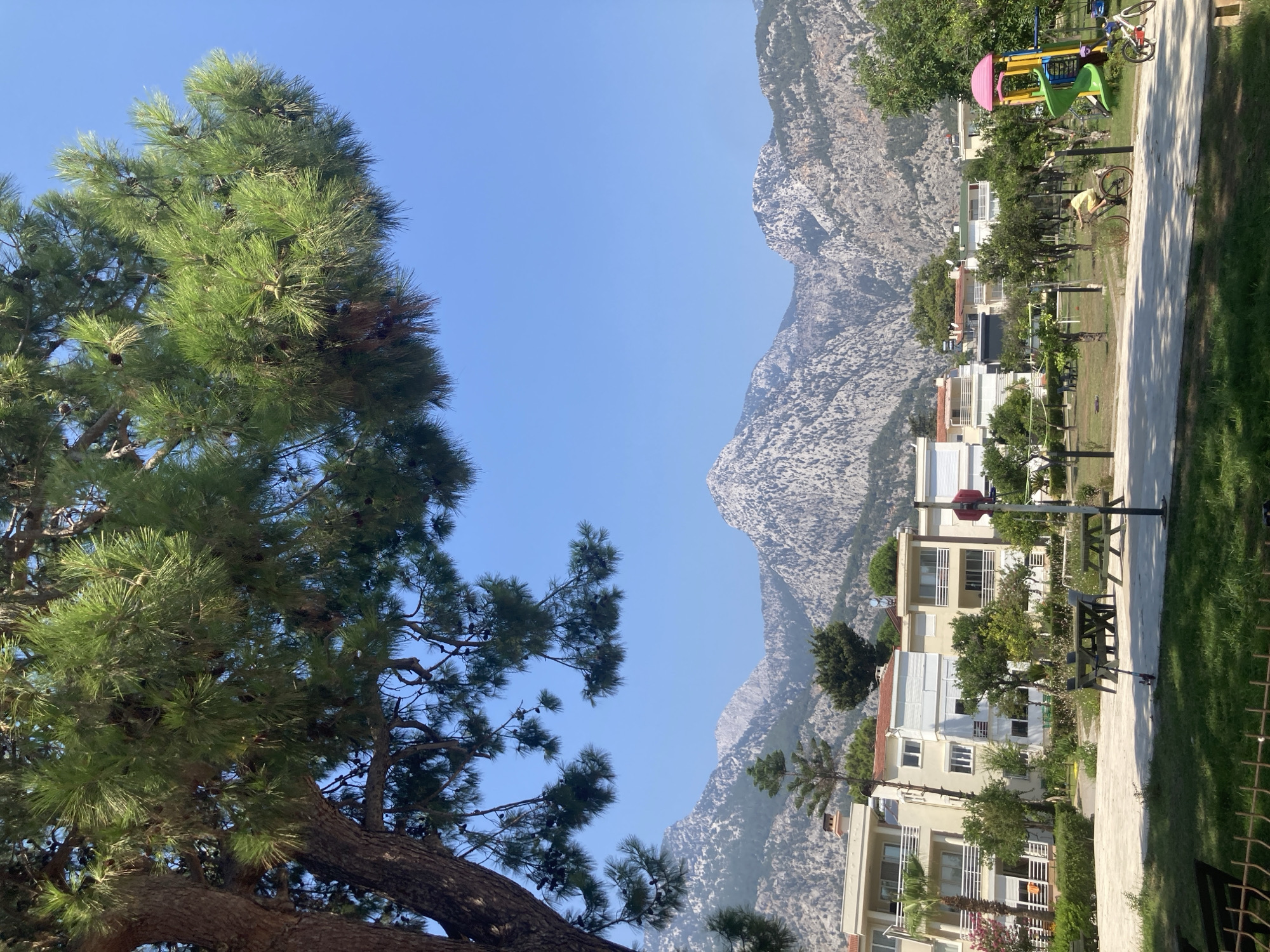 The image shows a view of residential buildings and a playground against a backdrop of rocky mountains under a clear blue sky. The foreground features a large pine tree with dense green foliage casting shadows. In the midground, there are several multi-story apartment buildings with balconies and white railings. A park area is visible with picnic tables, benches, and a children's playground structure with a green slide and yellow frame. Two children are present; one is riding a bicycle on a paved path, and another child is near a smaller bicycle. The mountains in the background are rugged and appear to be covered in sparse vegetation. The scene is consistent with the landscape found in Kemer, Turkey, known for its coastal location and proximity to mountainous terrain.